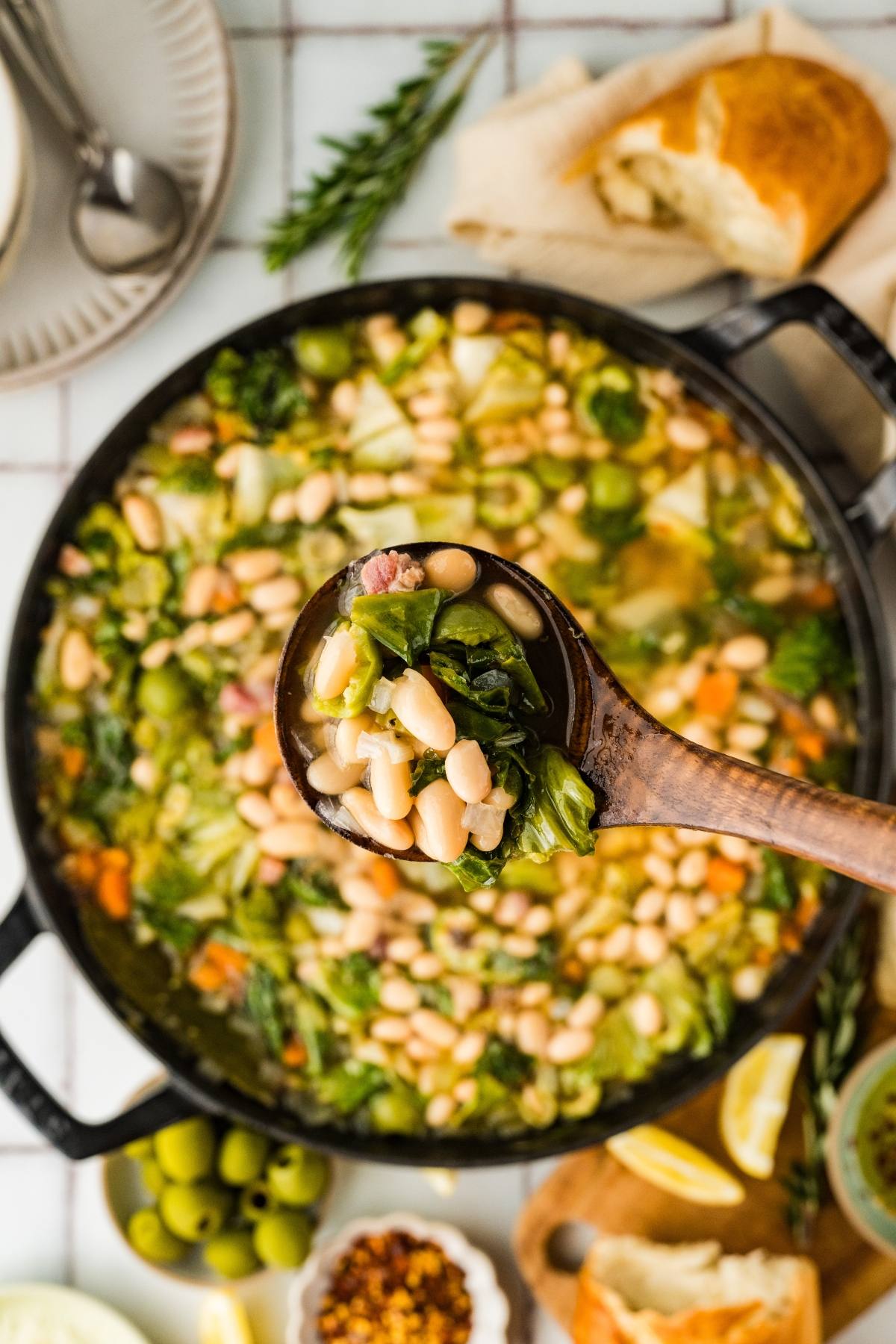 A ladle lifts white bean and greens soup from a pot, surrounded by bread, olives, and lemon wedges.