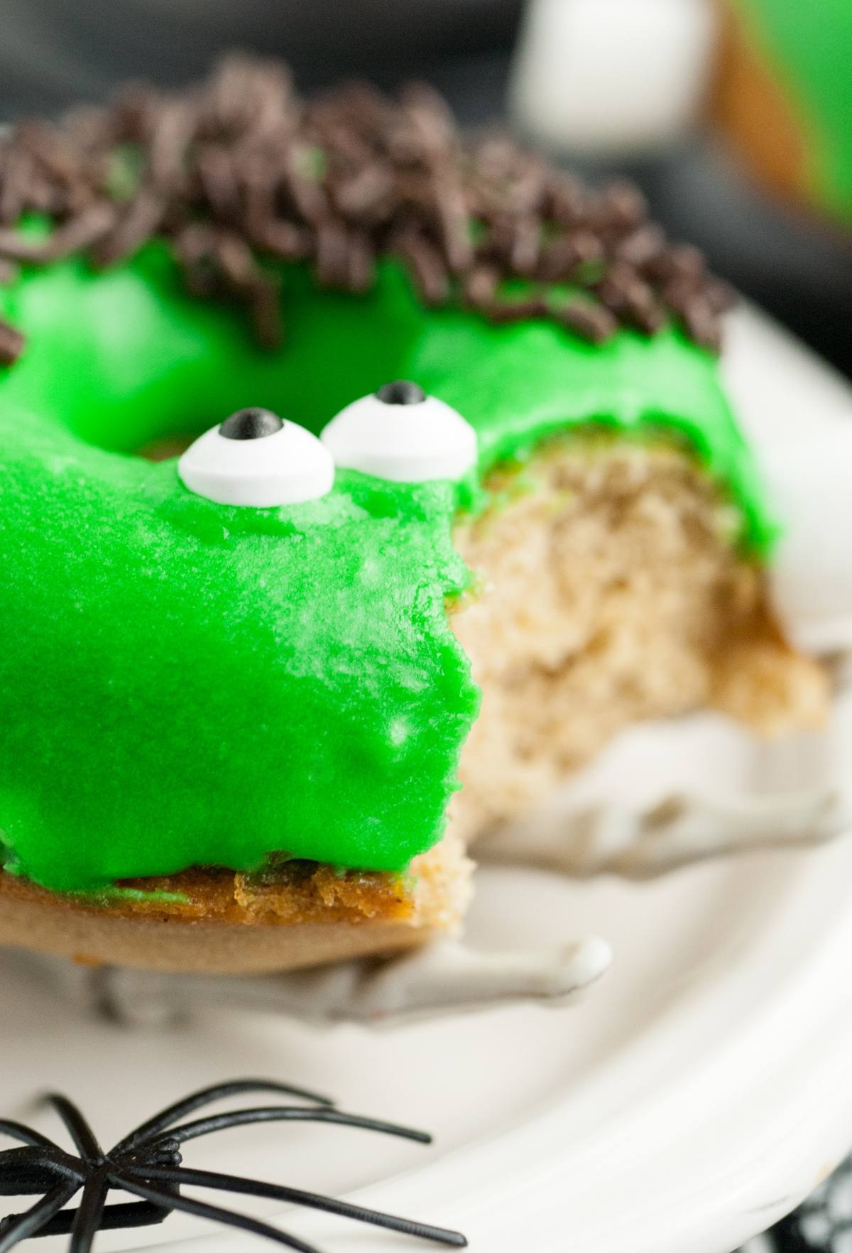 A spooky baked Halloween donut with green icing, candy eyes, and chocolate sprinkles, made to look like a Frankenstein monster—with a bite taken out!.