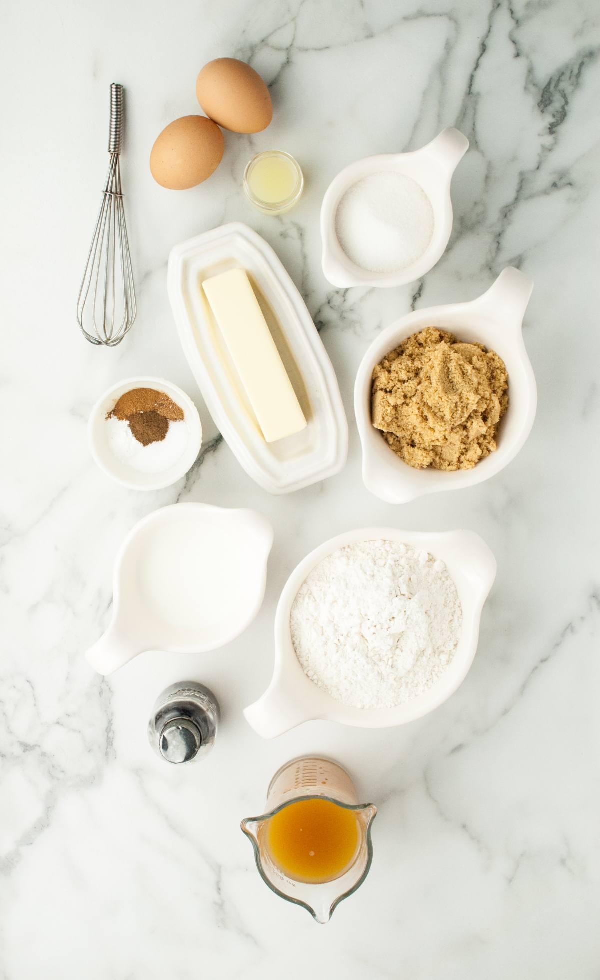 Baking ingredients on a marble surface: eggs, butter, sugars, flour, spices, milk, vanilla, whisk, and apple juice—perfect for Frankenstein Apple Cider Halloween Donuts that look like a Frankenstein monster.