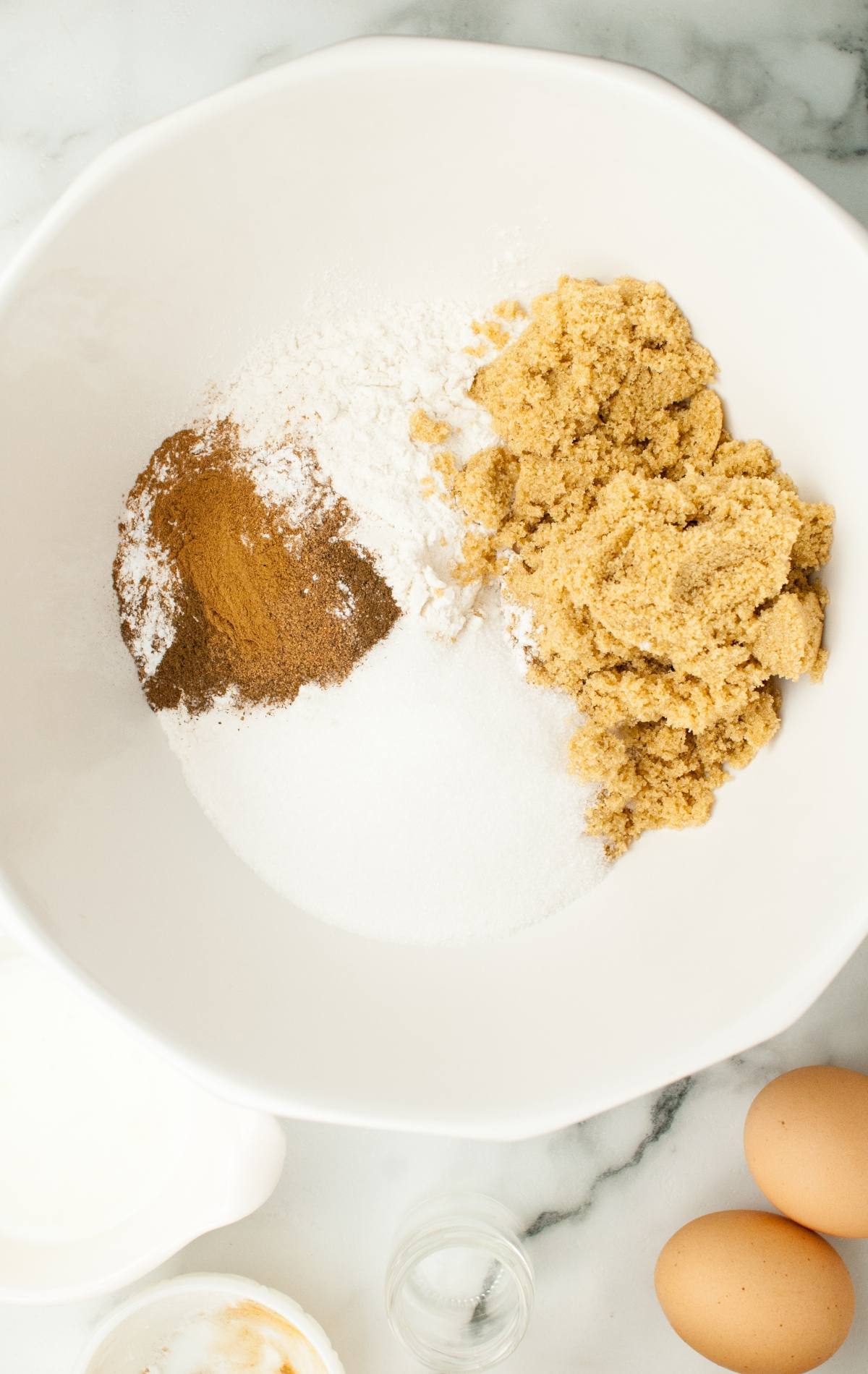 White bowl with brown sugar, white sugar, flour, and cinnamon sits beside eggs—perfect for making Frankenstein Apple Cider Halloween Donuts that look like a Frankenstein monster. These baked apple cider donuts are great for spooky season baking!.