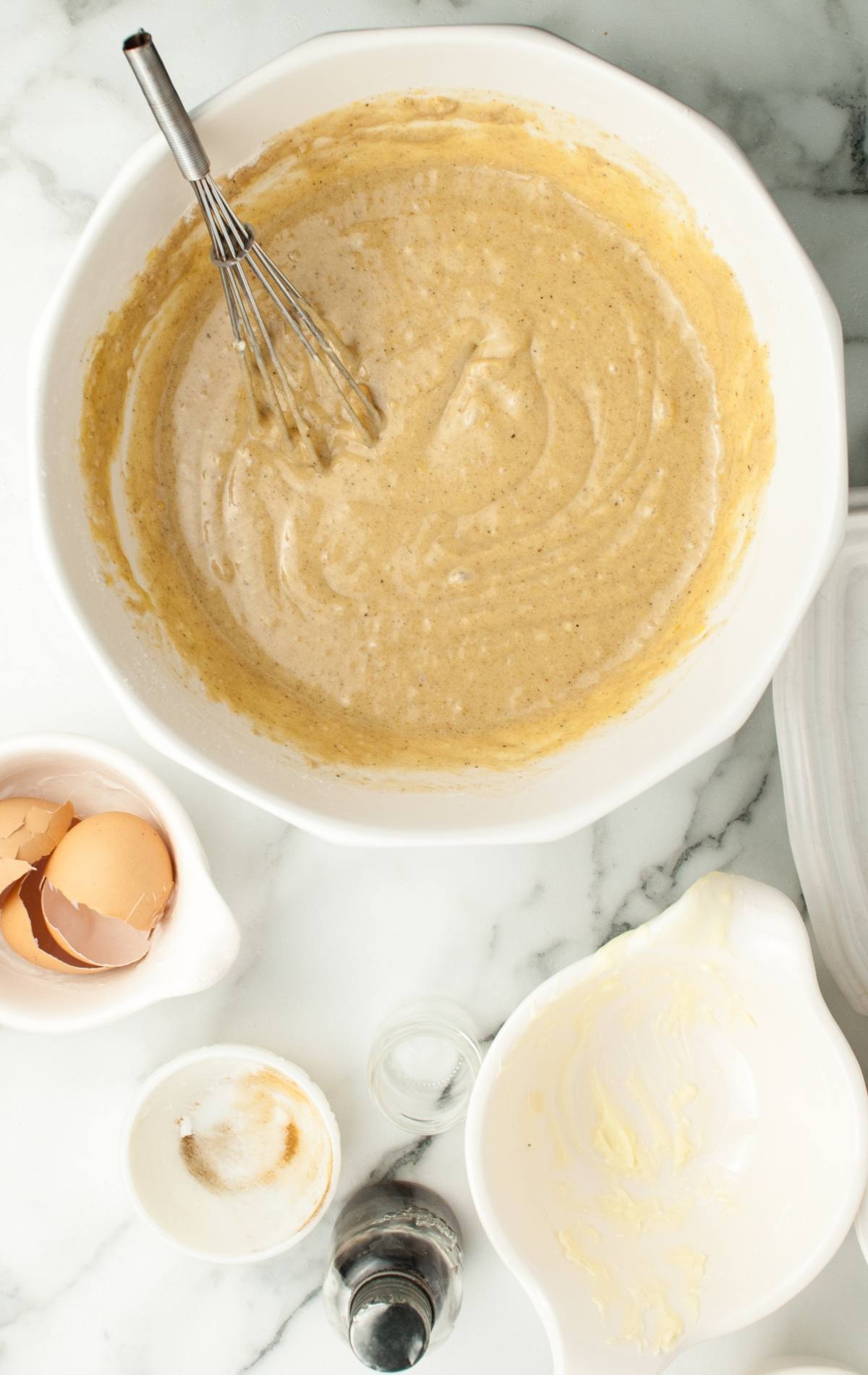 Whisk and banana bread batter in a bowl, with eggshells and ingredients on a marble countertop—looks like a Frankenstein monster ready to transform into something delicious.