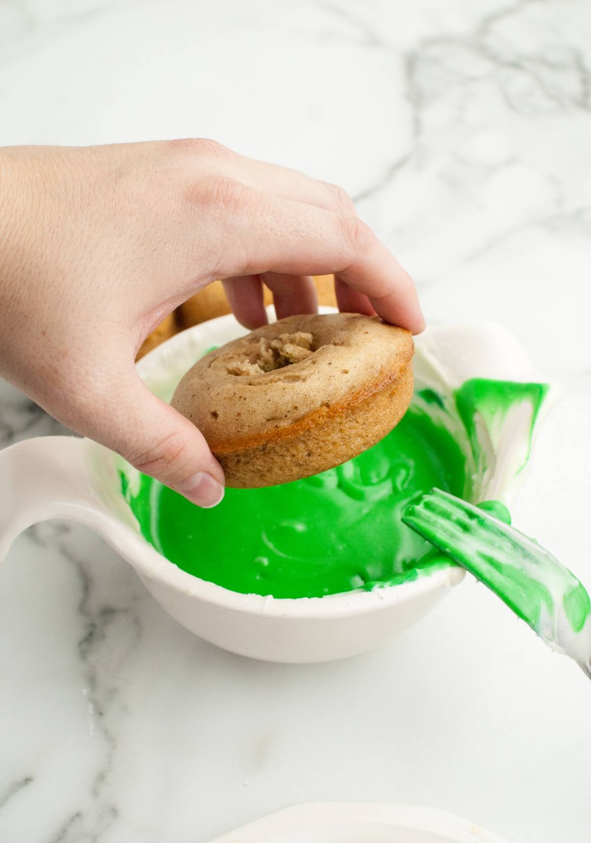 A hand dips a baked apple cider donut into a bowl of bright green icing on a marble countertop for some Halloween donut decorating.