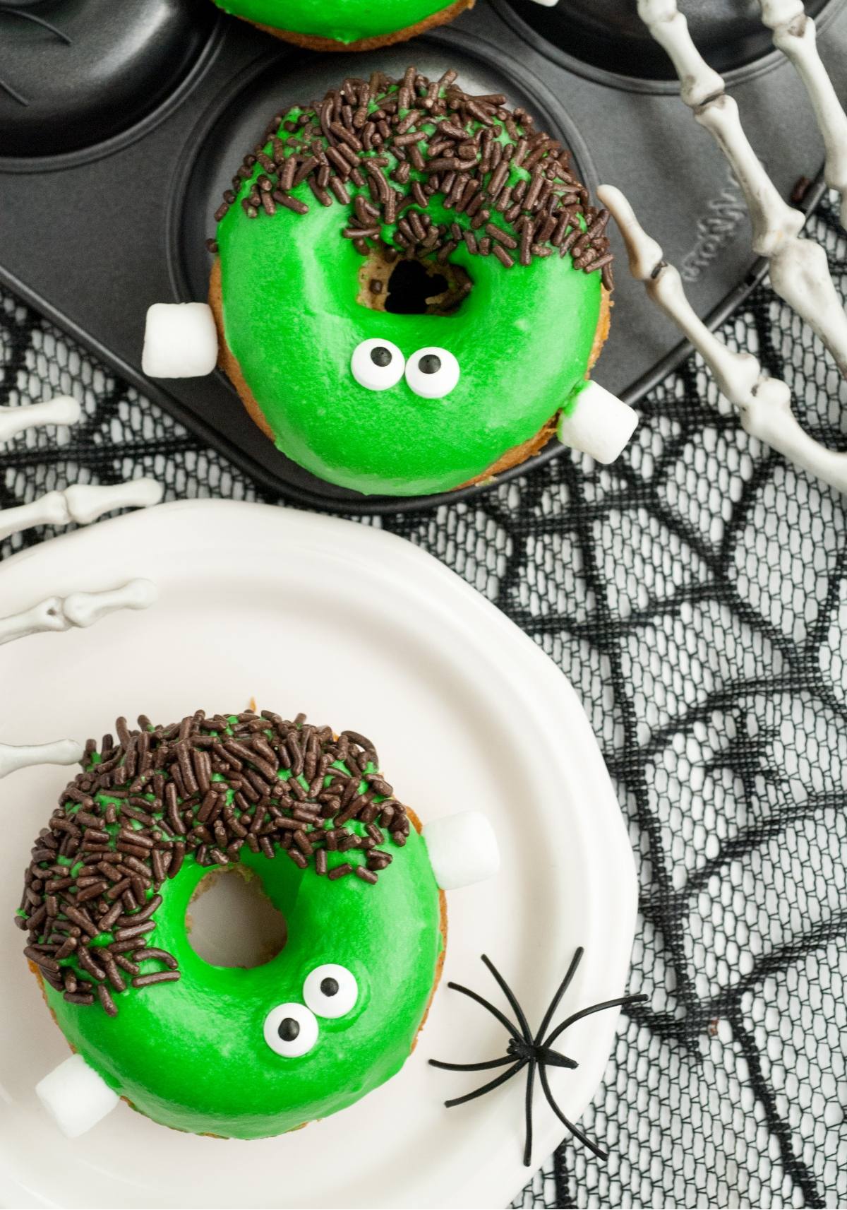 Green donuts decorated as Frankenstein's monster with candy eyes, chocolate sprinkles, and fake skeleton hands nearby—perfect for Halloween donut decorating or serving as Frankenstein Apple Cider Halloween Donuts.