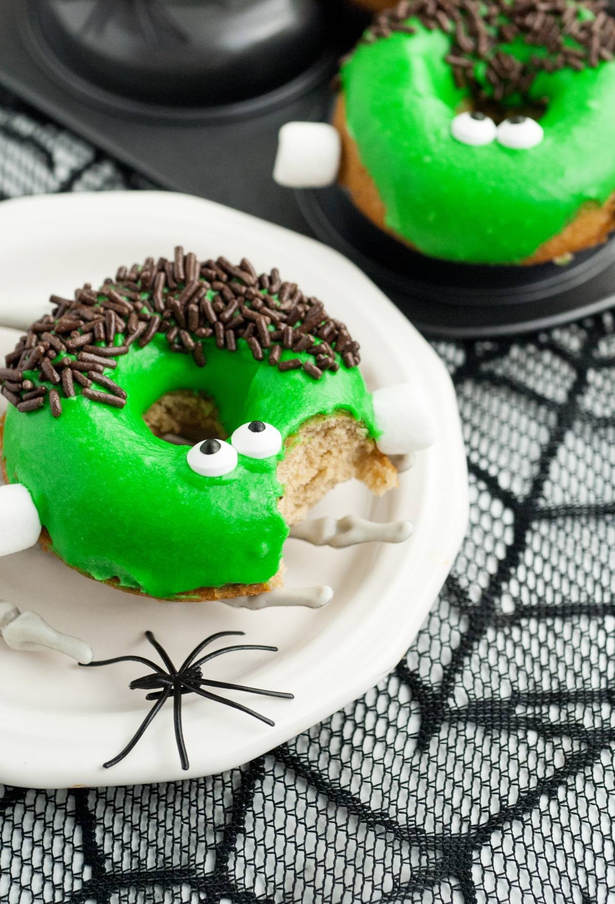 Green Spooky baked Halloween donut decorated like Frankenstein with candy eyes, sprinkles, and marshmallows on a spiderweb plate.