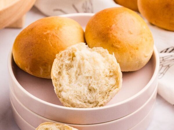 Three soft French bread rolls rest on a plate, one broken open to reveal its fluffy interior.