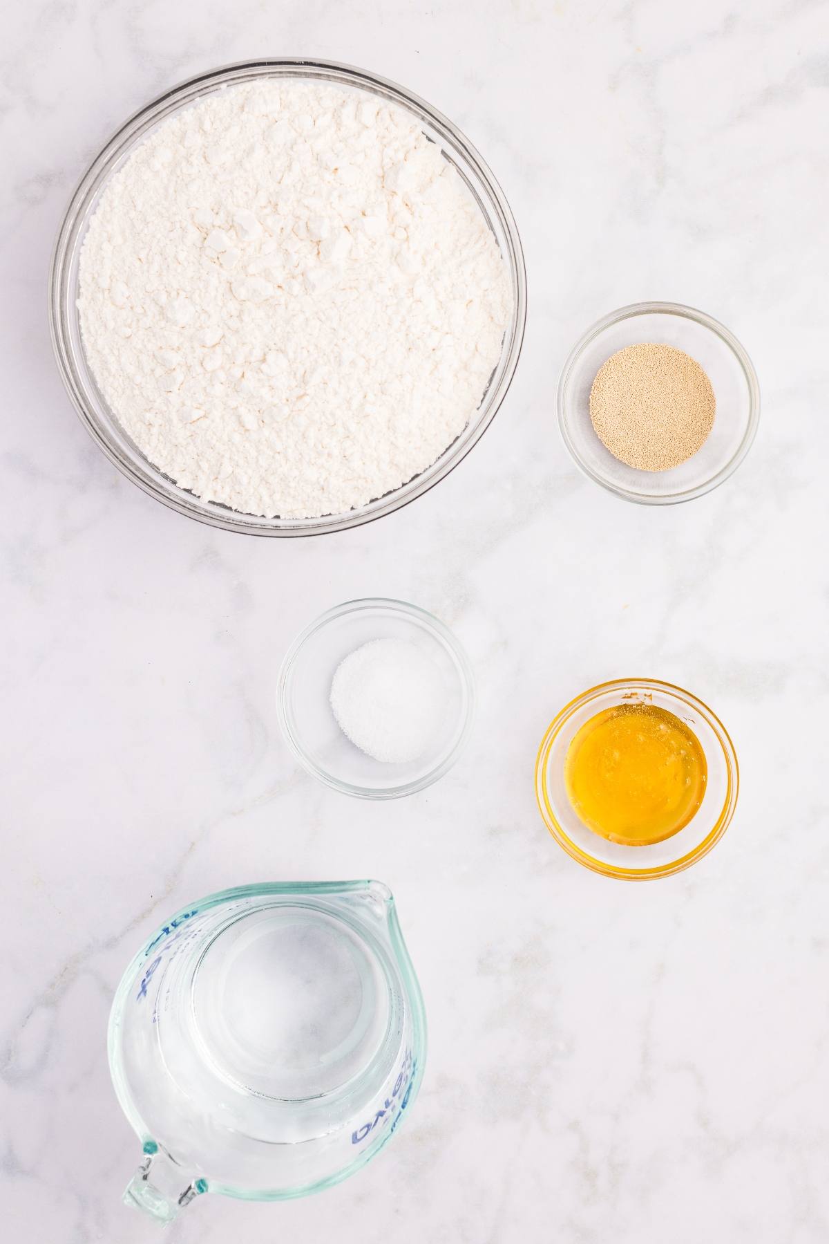 Five bowls with flour, yeast, salt, honey, and a measuring cup of water on a white marble surface.