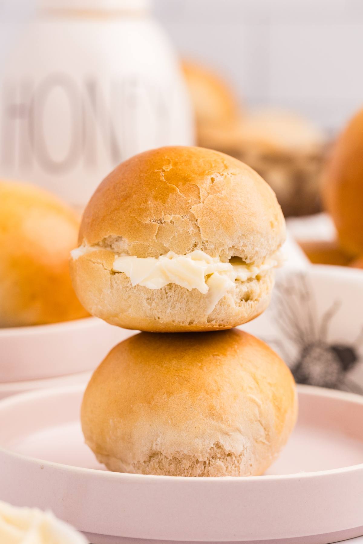 Two golden dinner rolls, one with butter filling, stacked on a pink plate with a honey jar in the background.