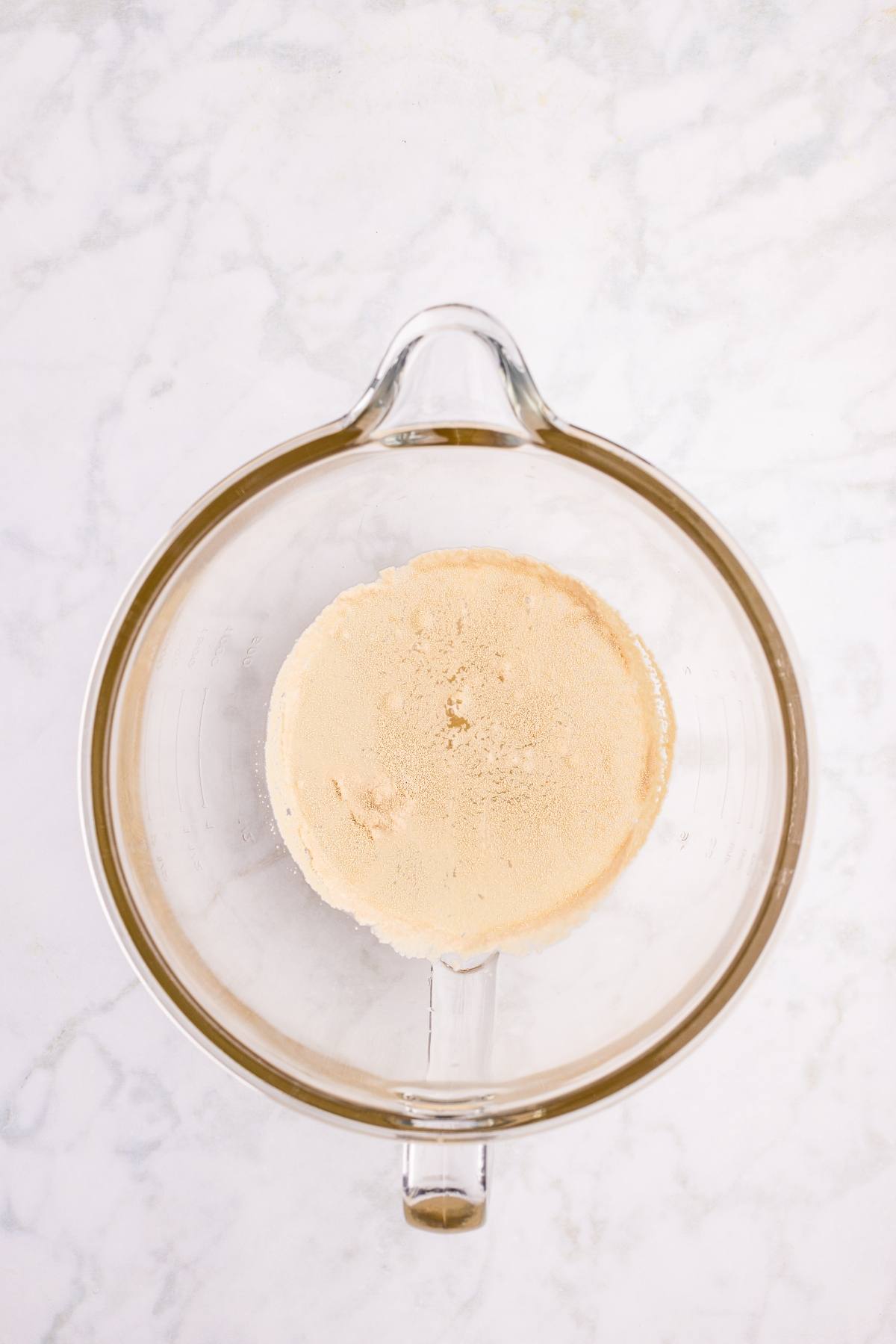 Overhead view of a glass mixing bowl with light brown dry ingredients on a white marble surface.