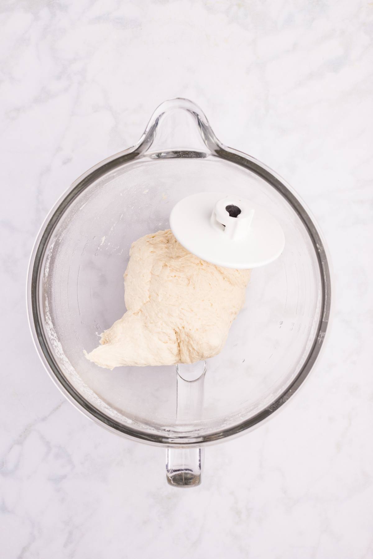 A glass mixing bowl with bread dough and a dough hook attachment on a light marble surface.