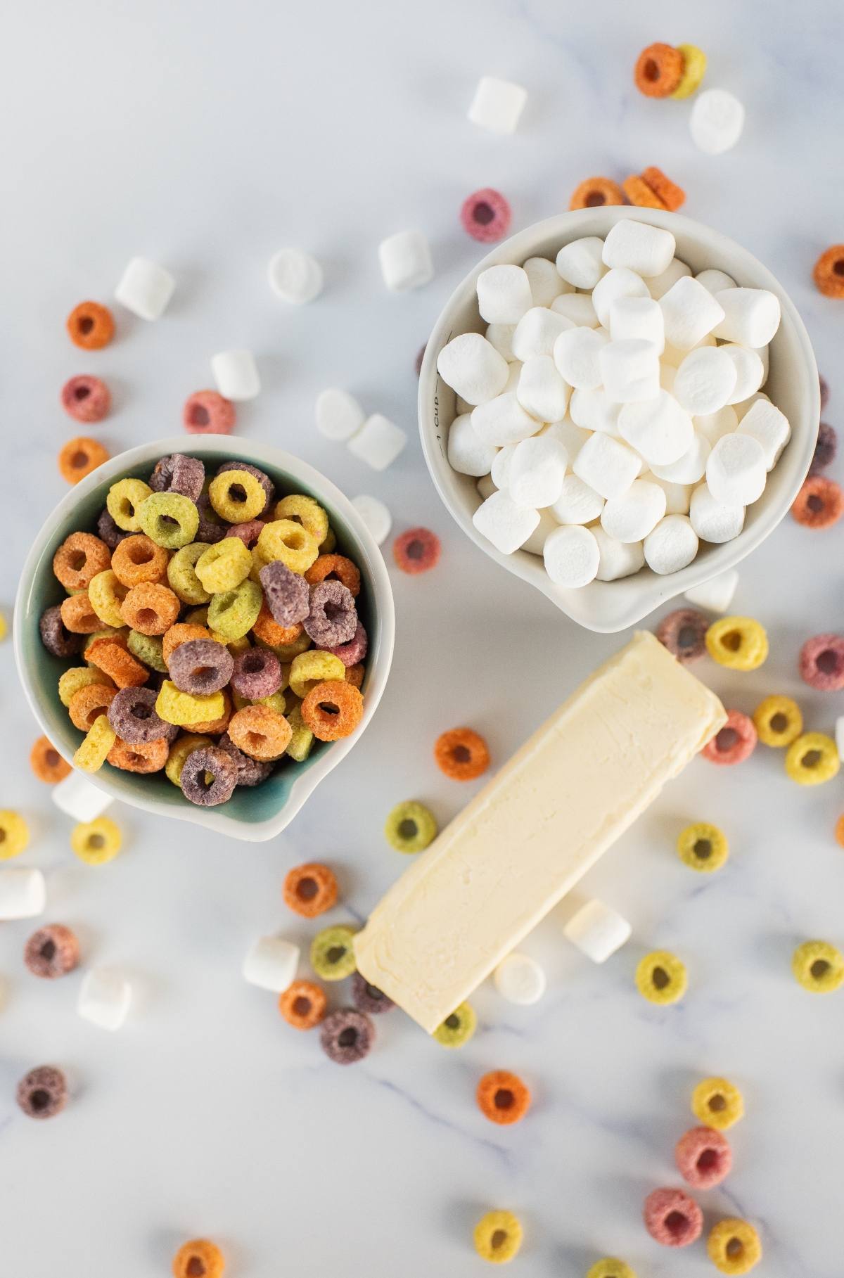 Two bowls with colorful cereal and mini marshmallows, a stick of butter, and scattered Fruit Loops Squares create a playful, vibrant scene.