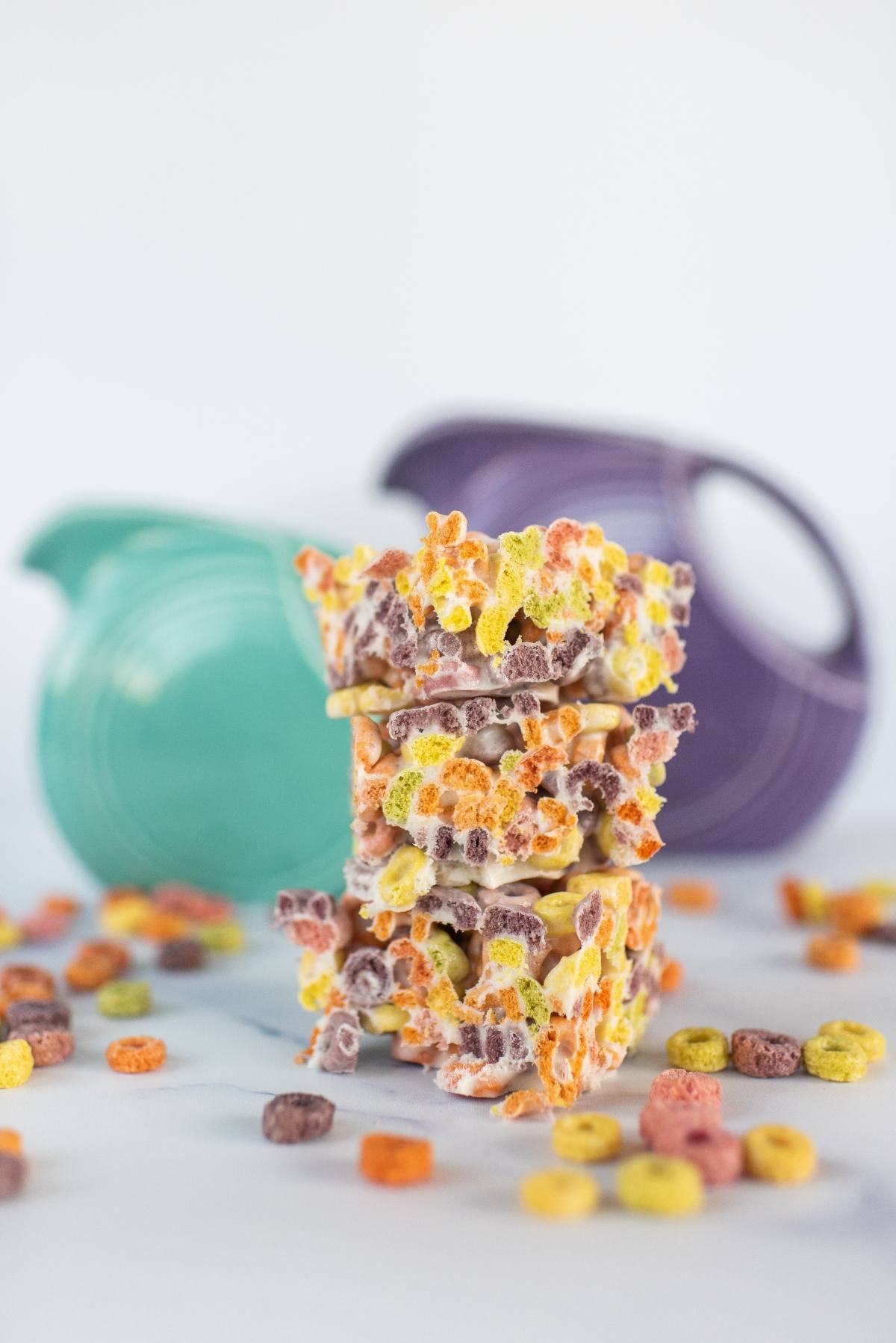 Three stacked fruit loops squares made of colorful cereal pieces with a blurred teal and purple pitcher in the background.