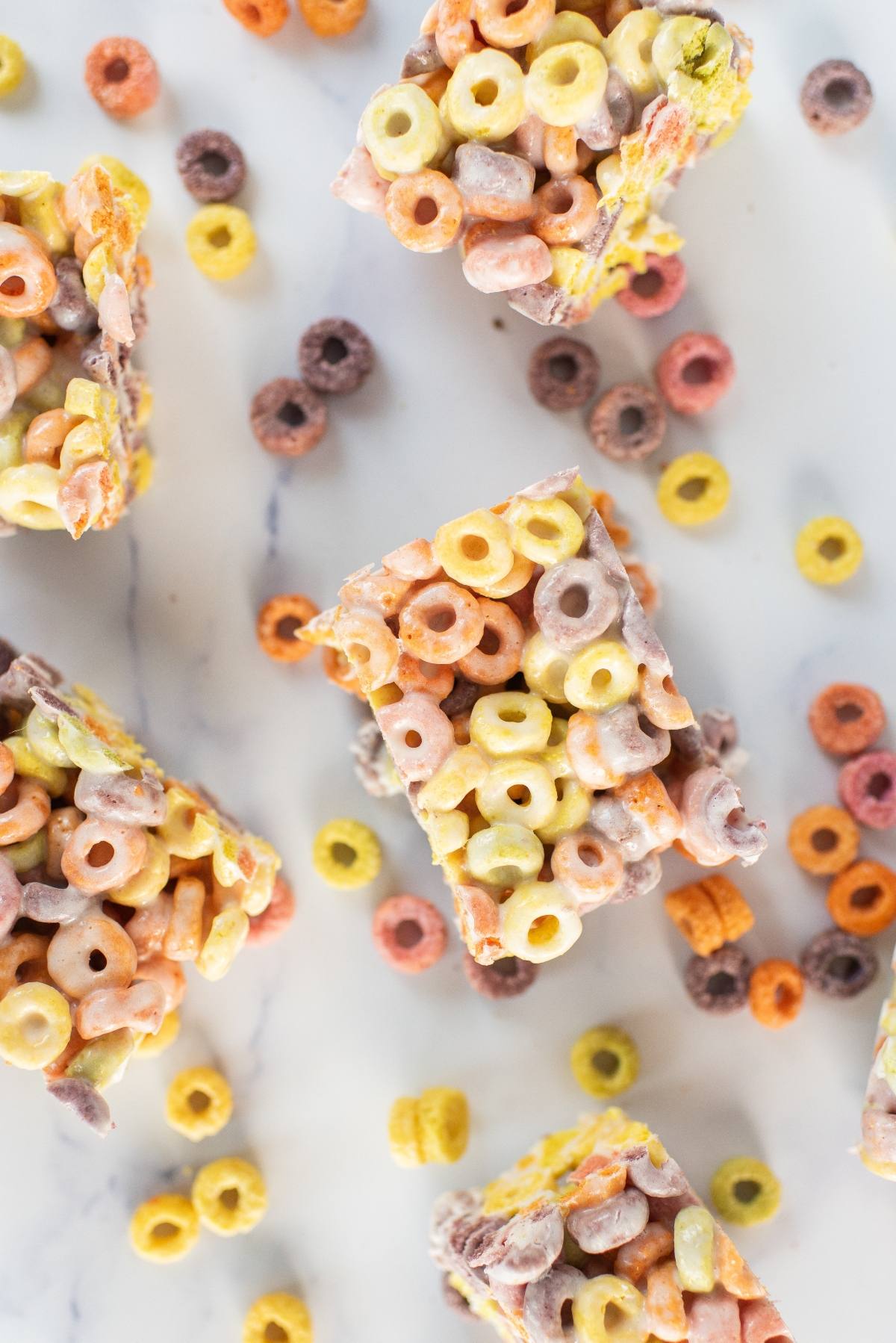 Colorful fruit loops squares and scattered multicolored cereal pieces rest on a white marble surface, viewed from above.