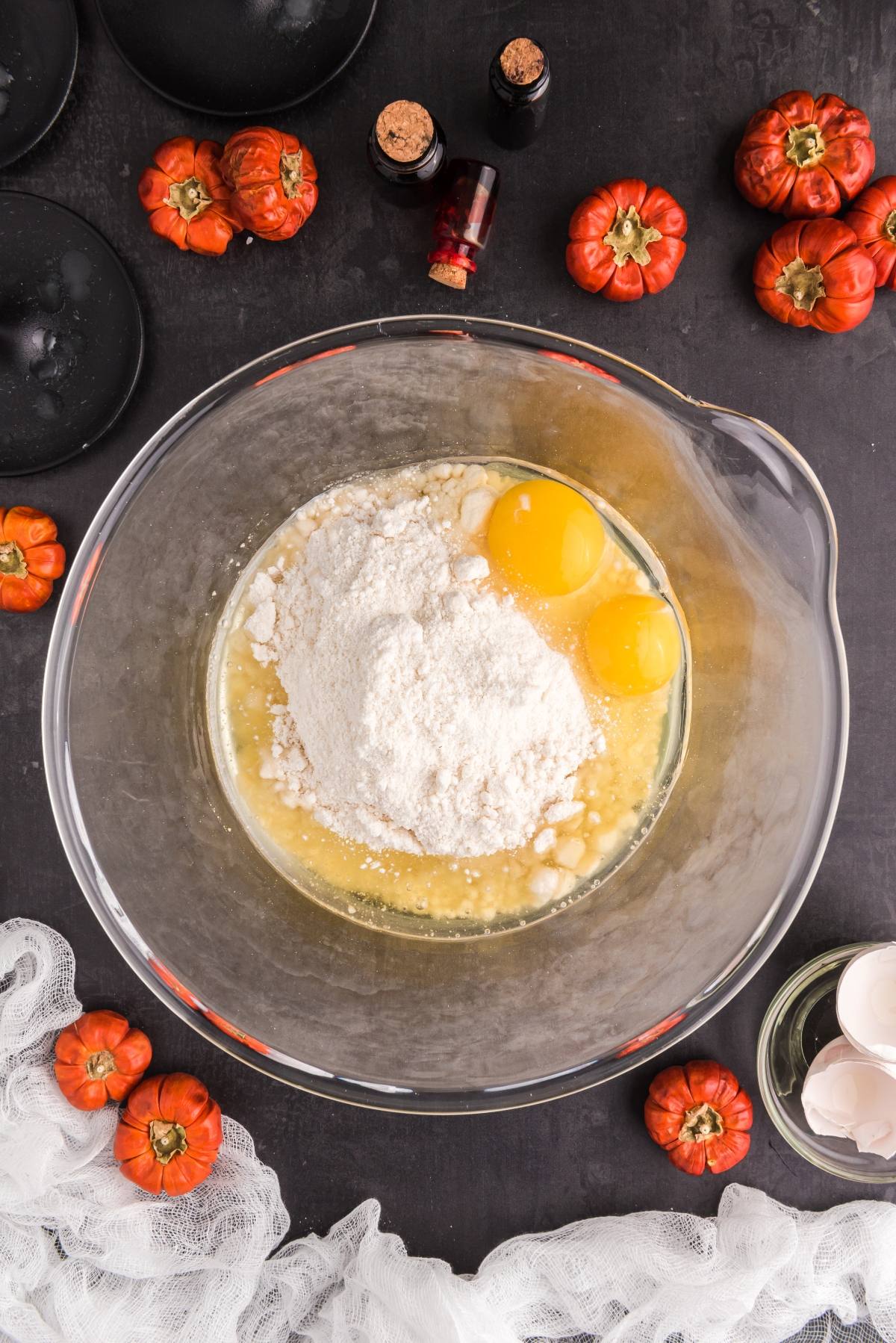 A glass bowl with flour, eggs, and liquid surrounded by mini pumpkins and baking ingredients on a dark surface&mdash;perfect for whipping up spooky Halloween treats like Halloween Crinkle Cookies.