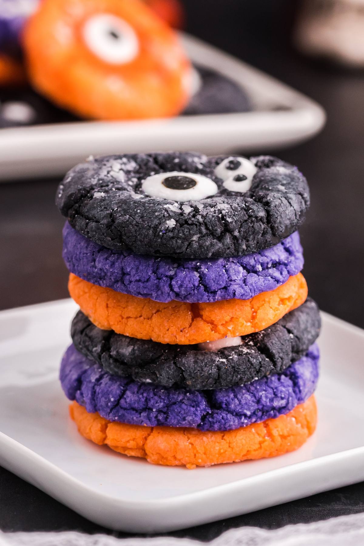 Stack of colourful monster cookies in orange, purple, and black with candy eyes on a white plate&mdash;perfect for easy Halloween cookie decorating ideas.