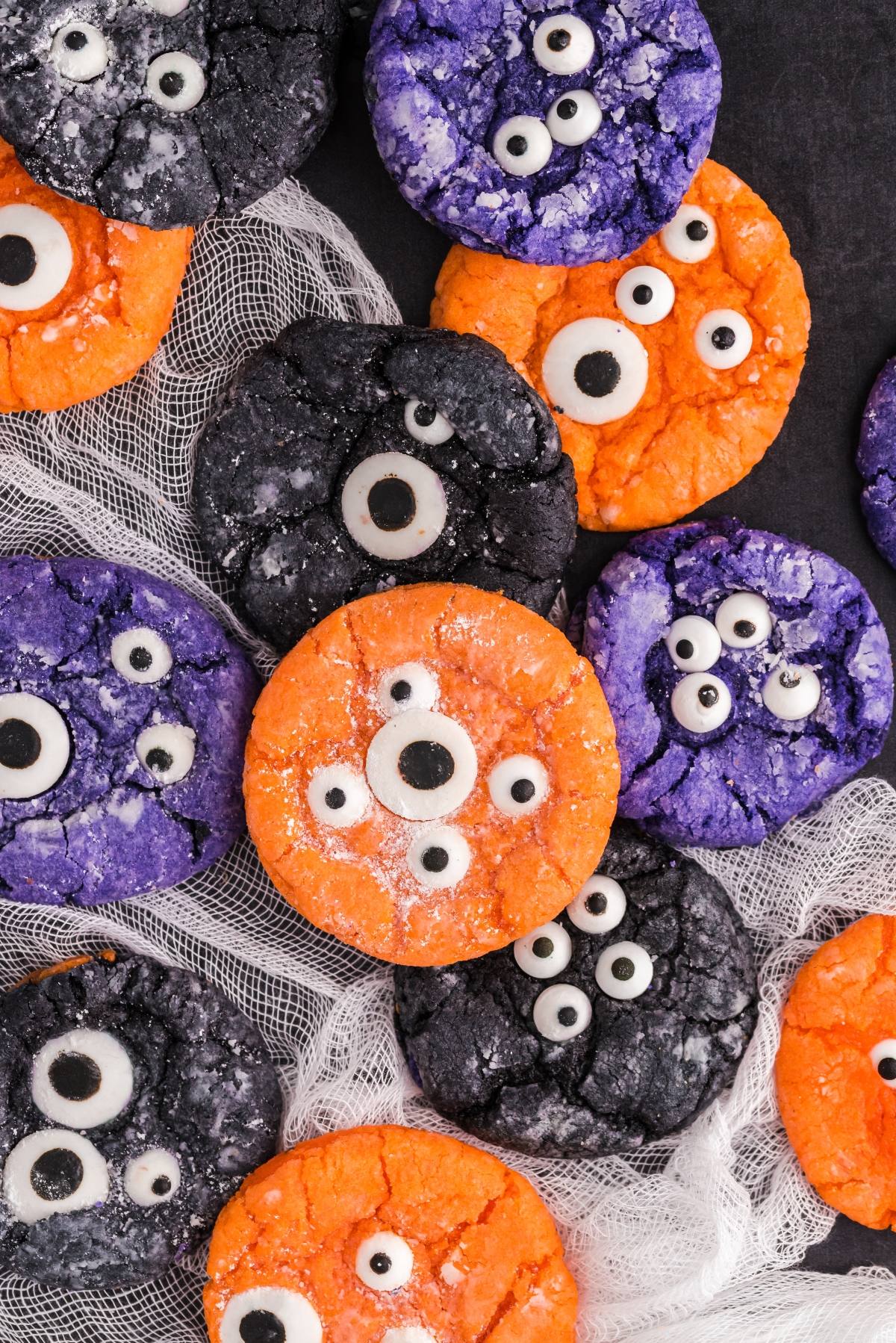 Colorful monster cookies in orange, purple, and black, decorated with candy eyeballs, are arranged on a white netted fabric&mdash;perfect Halloween Crinkle Cookies for your spooky celebration.