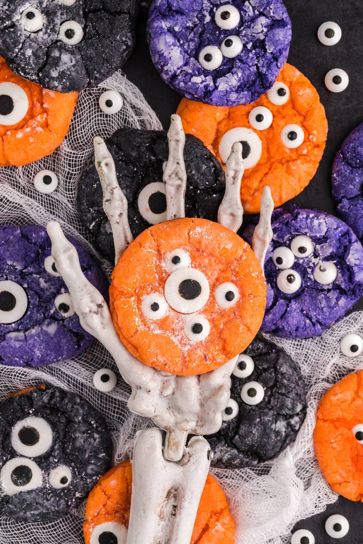 A skeleton hand holds an orange monster cookie with candy eyes, surrounded by purple and black cake mix Halloween cookies&mdash;perfect for easy Halloween cookie decorating ideas.