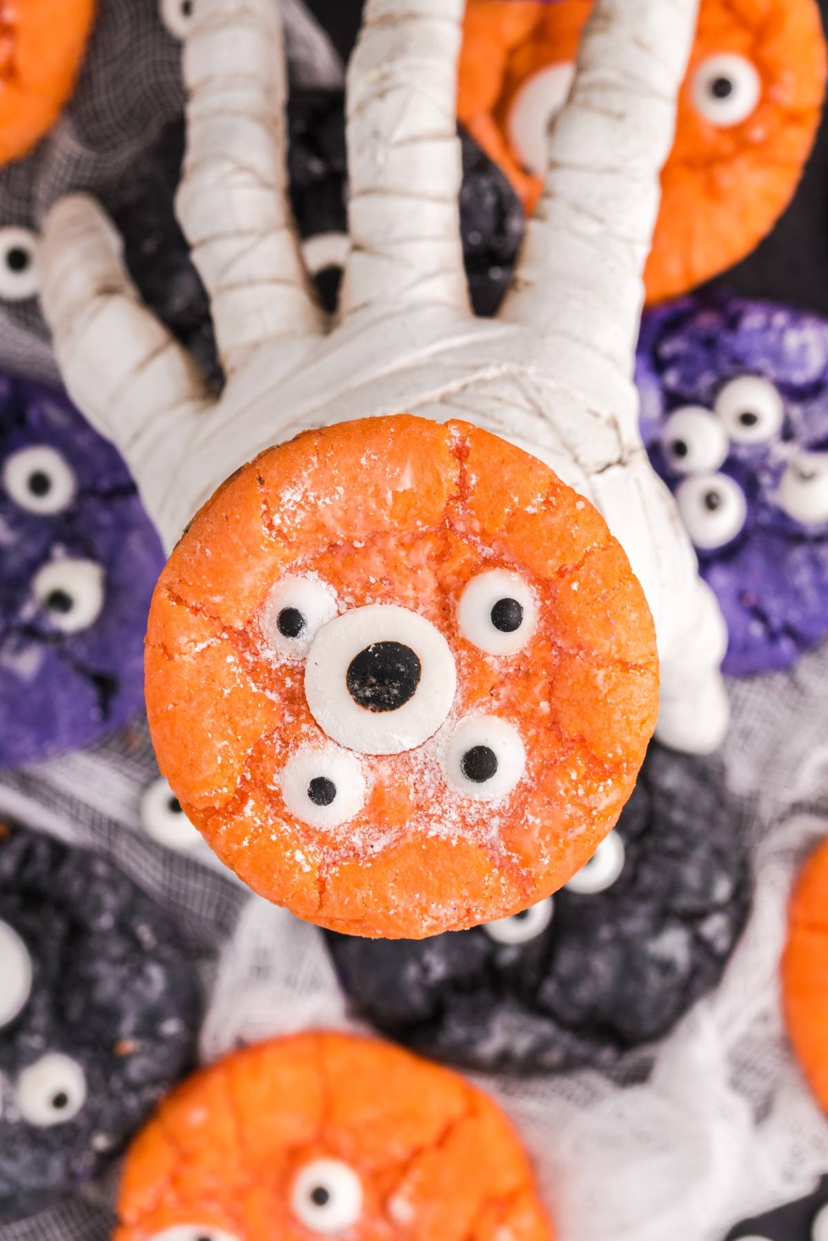 A hand in white gauze holds an orange cookie with candy eyes; below are colourful monster cookies, perfect as Halloween Crinkle Cookies made from cake mix.