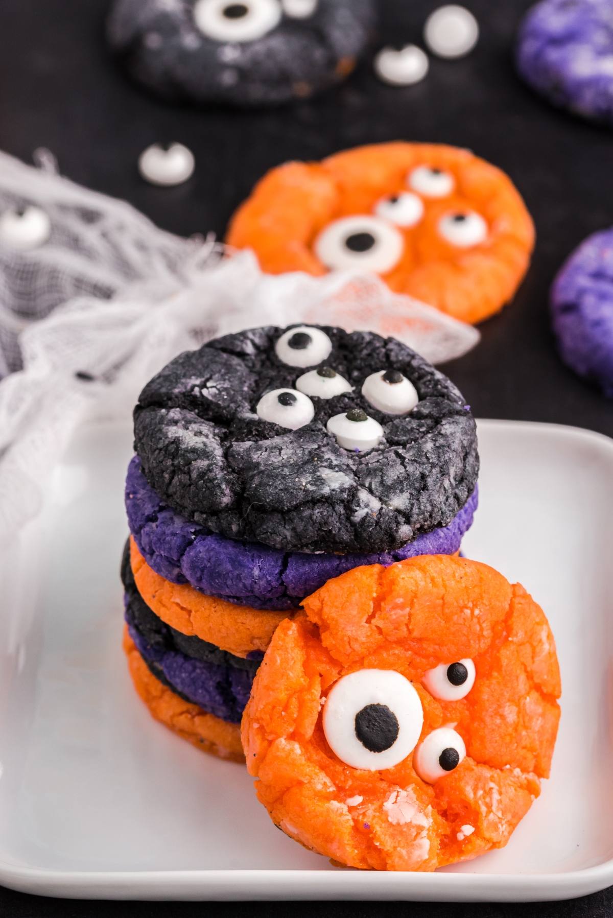 A stack of colourful monster cookies with candy eyes, decorated in orange, purple, and black on a white plate&mdash;spooky Halloween treats perfect for any festive gathering.