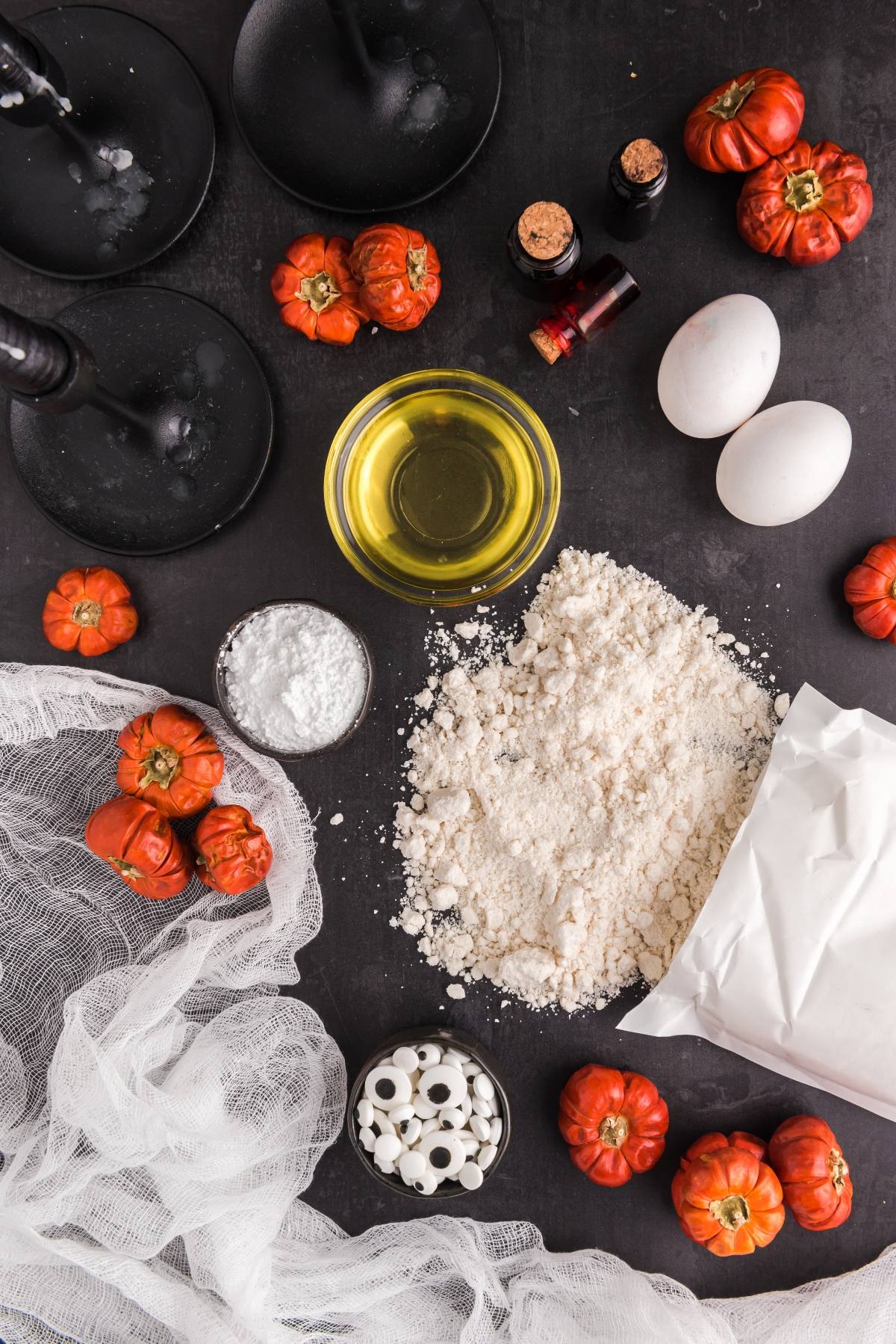 Baking ingredients, eggs, oil, flour, and Halloween decorations sit on a dark table with fake gauze and mini pumpkins&mdash;perfect for making spooky Cake mix Halloween cookies or Colourful monster cookies.