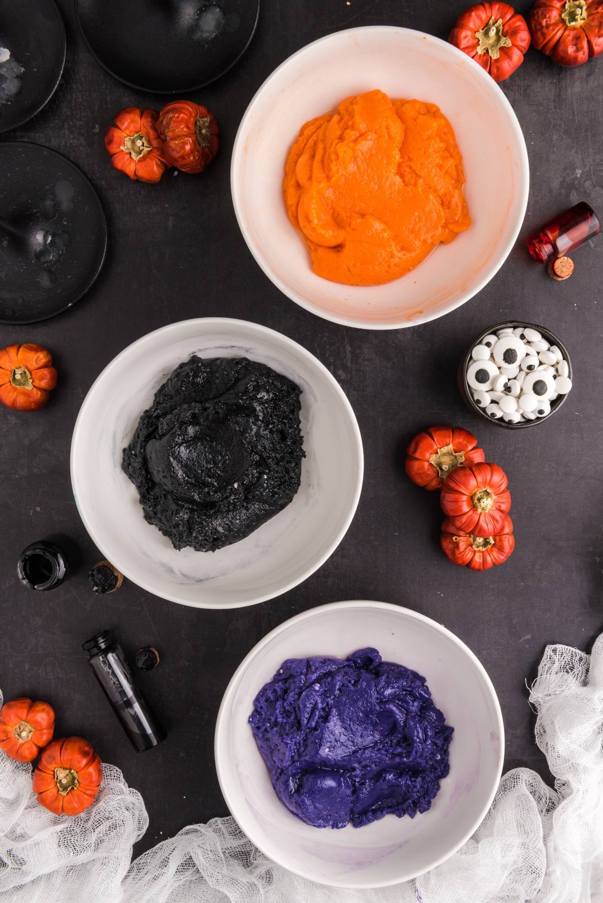 Three bowls filled with vibrant orange, black, and purple frosting&mdash;perfect for colourful monster cookies or easy Halloween cookie decorating ideas&mdash;are surrounded by mini pumpkins and candy eyeballs.