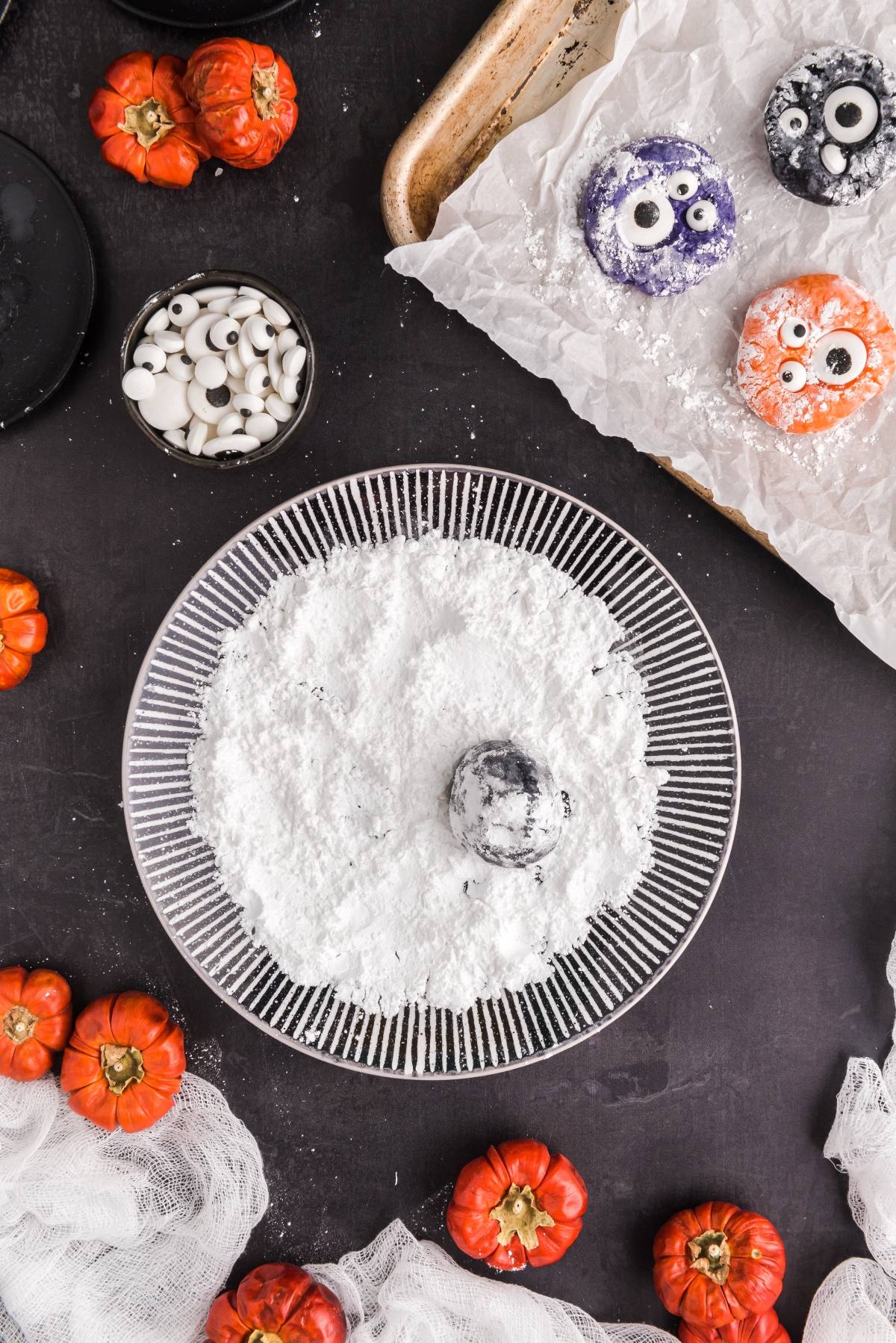 A plate of powdered sugar with colorful monster donuts and candy eyes, surrounded by mini pumpkins&mdash;perfect alongside spooky Halloween treats like Halloween Crinkle Cookies.