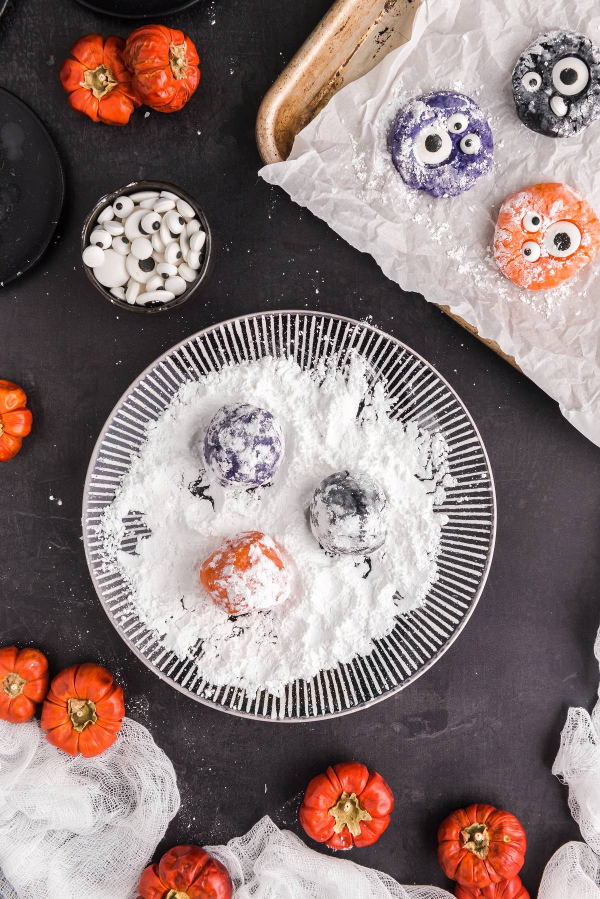 Three colorful Halloween Crinkle Cookies dusted with powdered sugar sit on a plate, featuring candy eyes and mini pumpkins&mdash;a fun display for easy Halloween cookie decorating ideas.