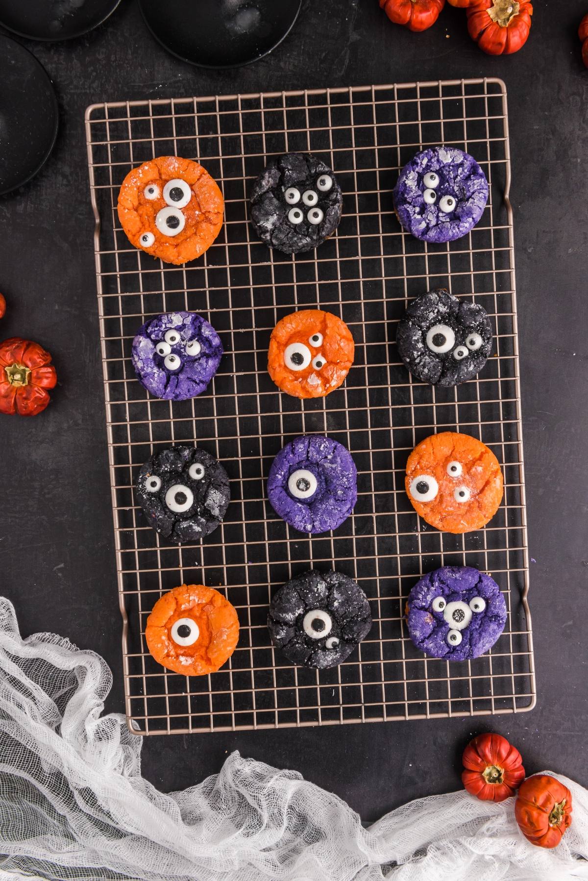 Colorful Halloween monster cookies with candy eyes on a cooling rack, surrounded by mini pumpkins&mdash;these spooky Halloween treats are perfect for your next party. Try making them as Halloween Crinkle Cookies for extra fun!.
