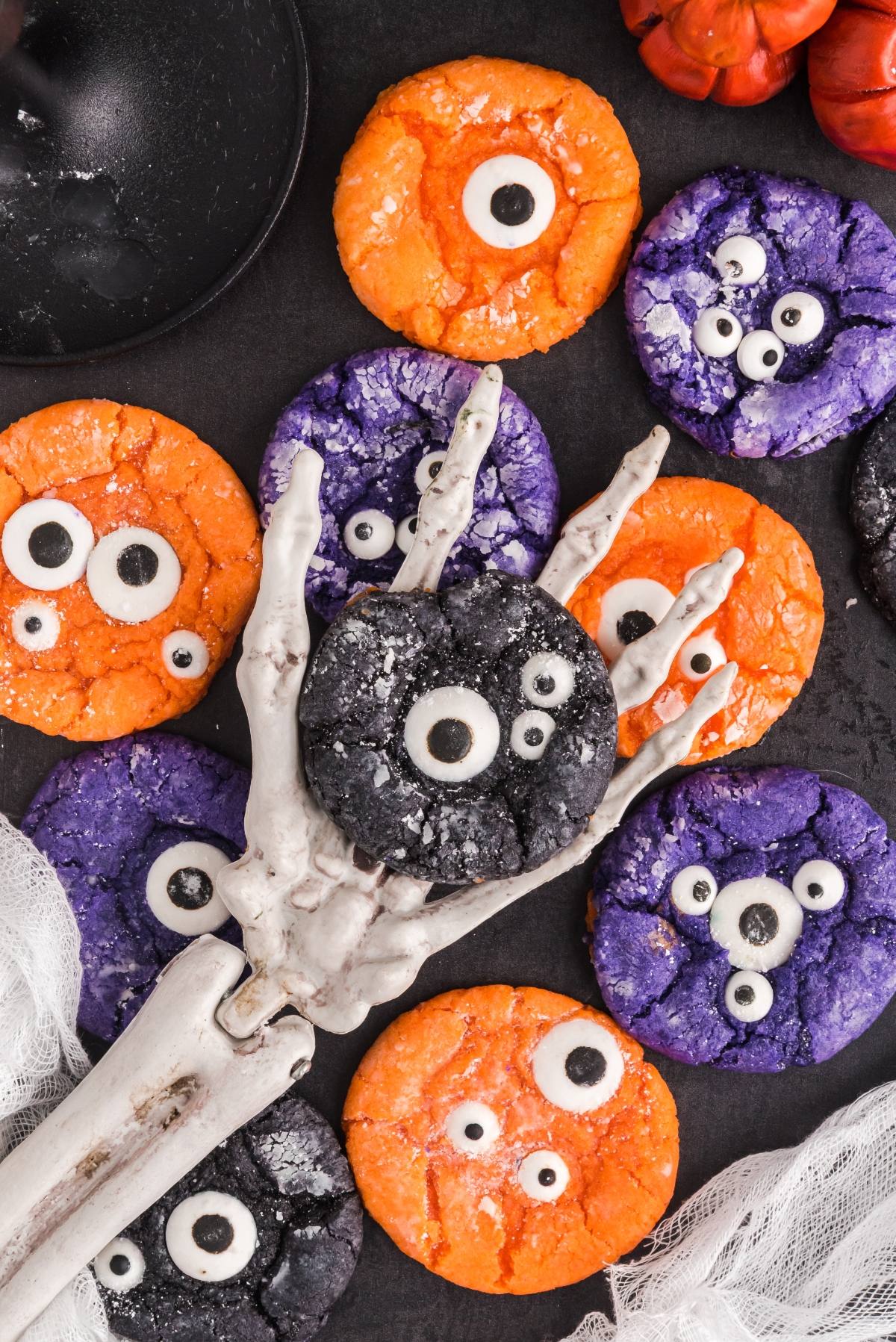 Colorful monster cookies with candy eyes, made using cake mix Halloween cookies recipes, are held by a skeleton hand and surrounded by white fabric and pumpkins&mdash;perfect for easy Halloween cookie decorating ideas.