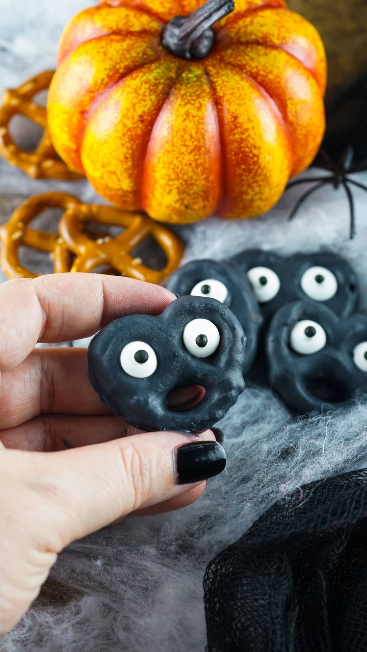 Hand holding a black Halloween Monster Eye Pretzel with a spooky face and candy eyes; pumpkin and pretzels in the background.
