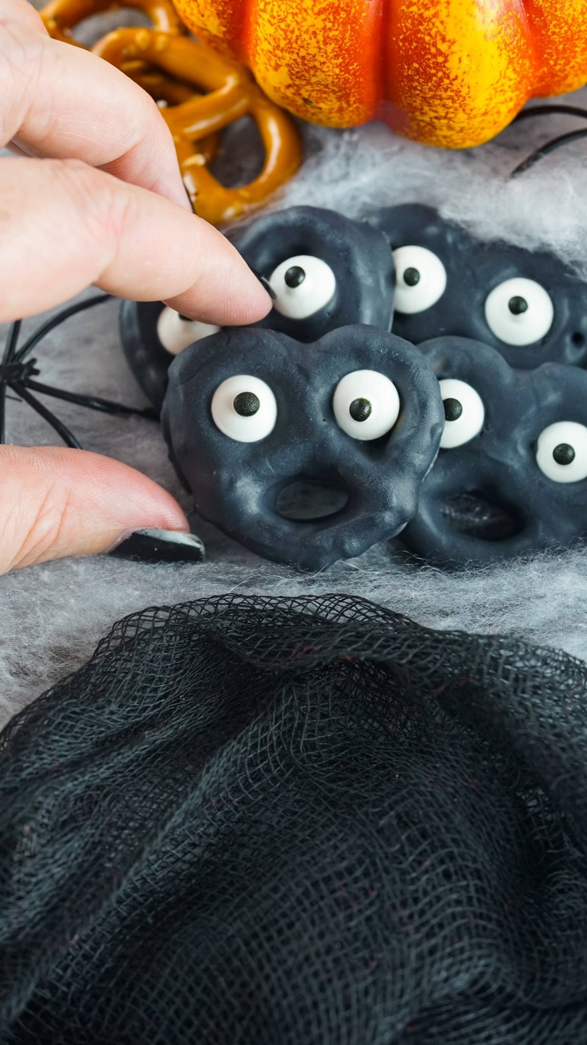 A hand picks up a black Halloween Monster Eye Pretzel decorated with candy eyes, surrounded by more spooky pretzels and Halloween props—an easy party treat recipe perfect as a Halloween snack for kids.
