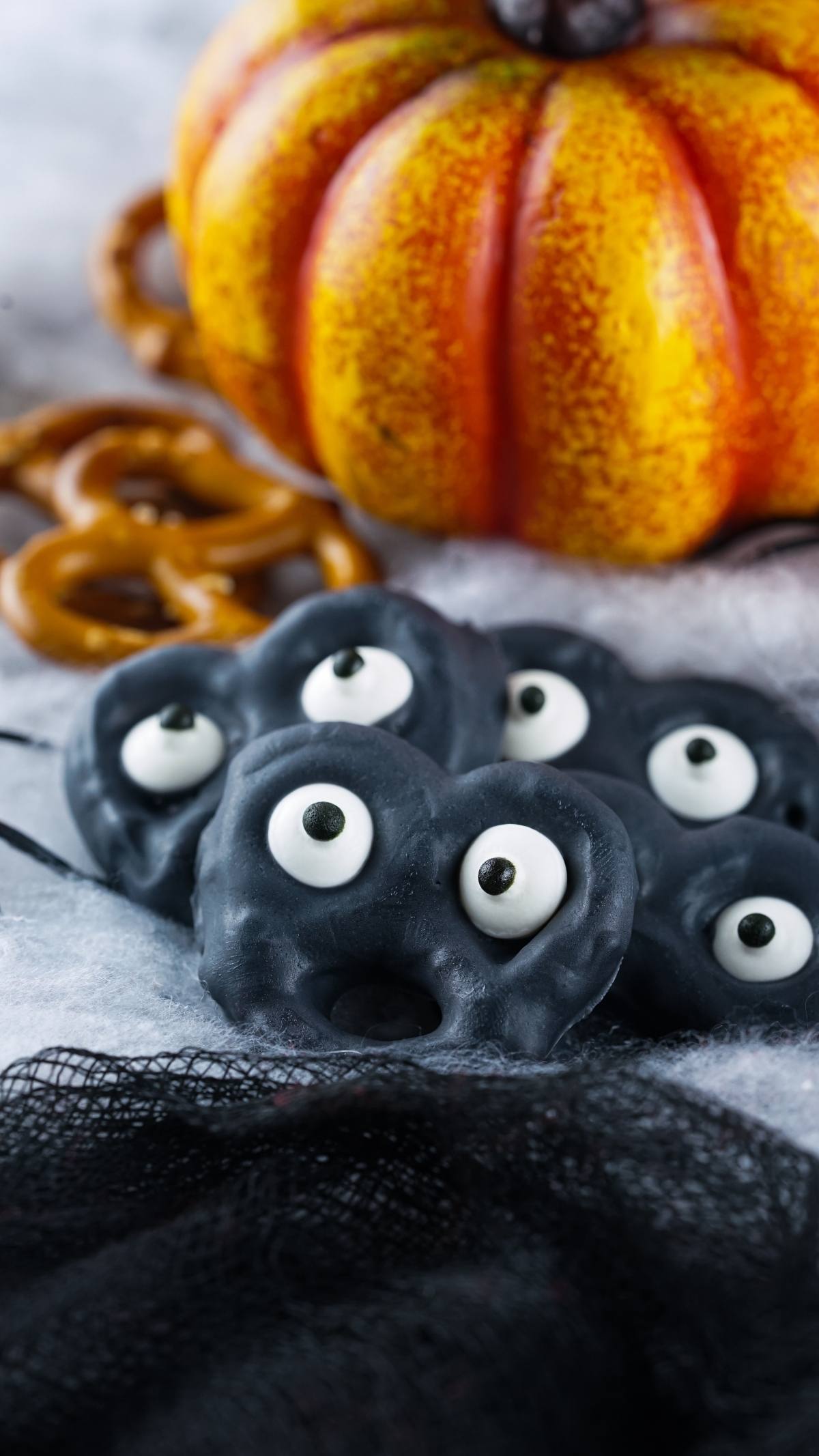 Black pretzels with googly eyes, a fake pumpkin, and brown pretzels on a fuzzy white surface create a fun Halloween snack for kids.