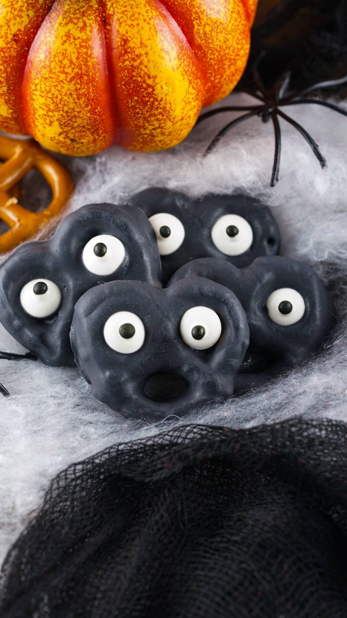 Four black Halloween pretzels with eyeballs are arranged on cobwebs, with spiders and a pumpkin in the background—an easy party treat recipe and a perfect Halloween snack for kids.