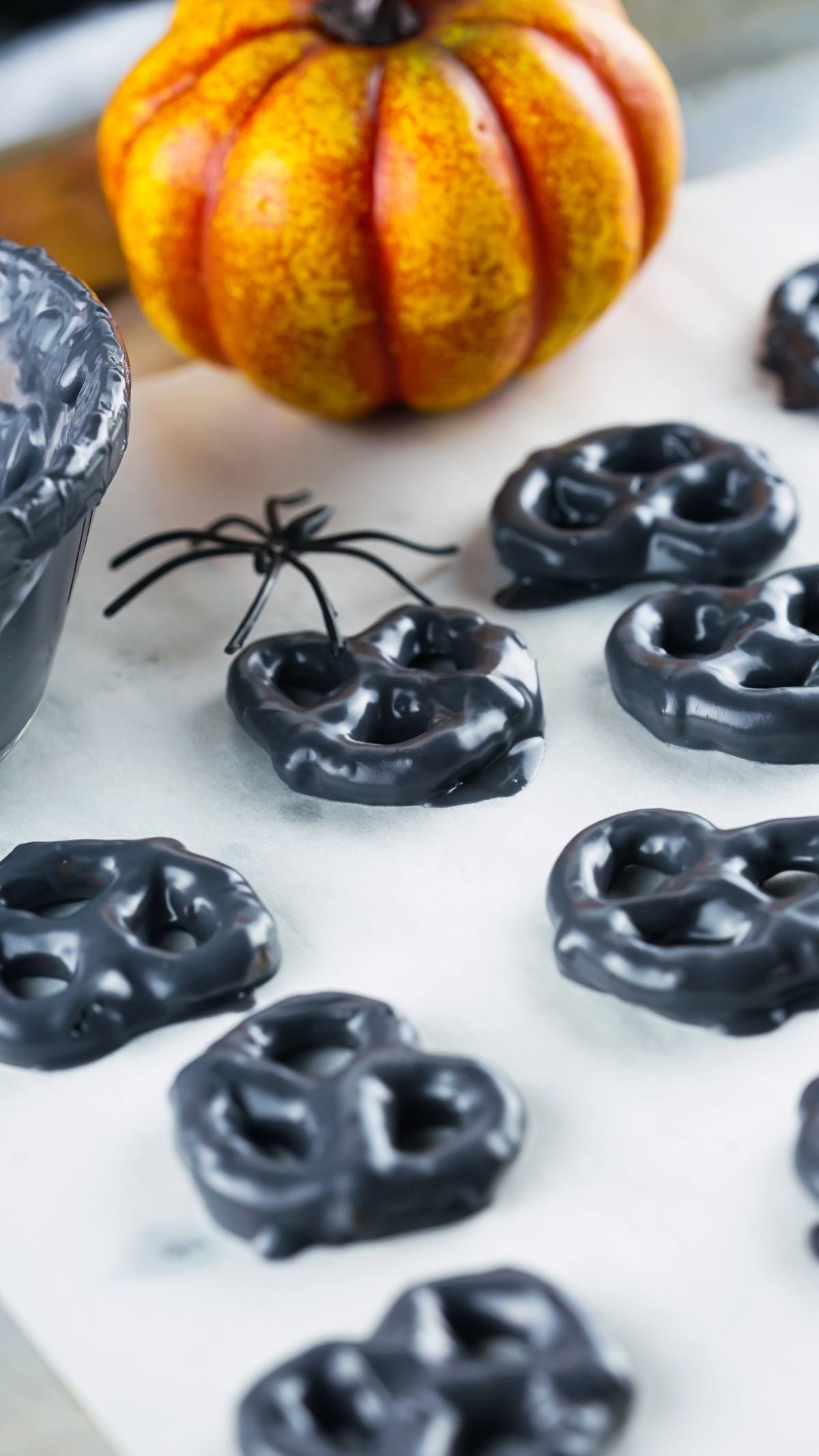 Black chocolate-covered pretzels on parchment make an easy party treat recipe, perfect for Halloween. With a fake spider and pumpkin in the background, these spooky monster treats are sure to be a hit!.