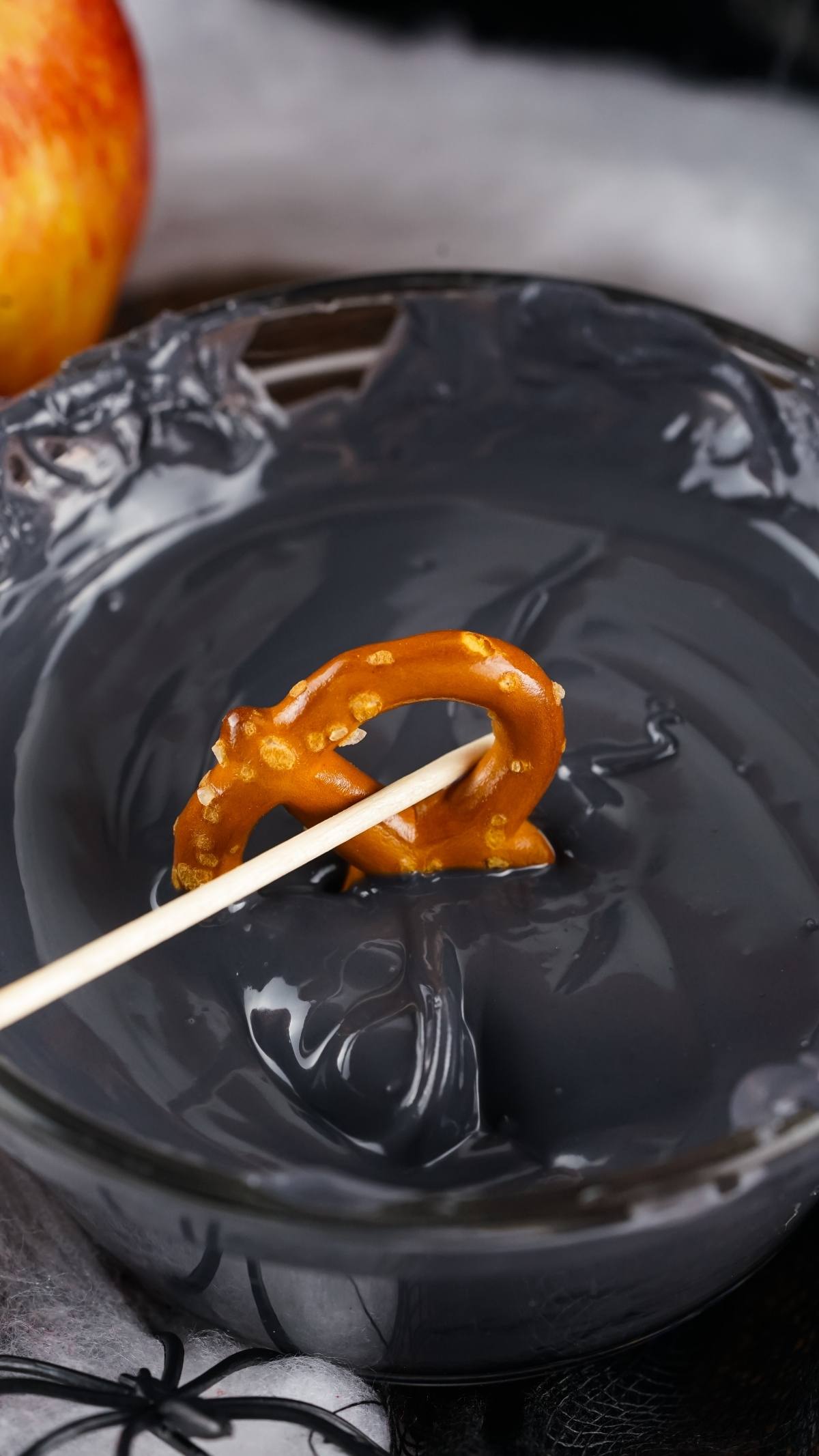 A pretzel on a stick is dipped into a bowl of glossy black liquid, perfect for making Halloween Monster Eye Pretzels—a spooky and fun Halloween snack for kids.