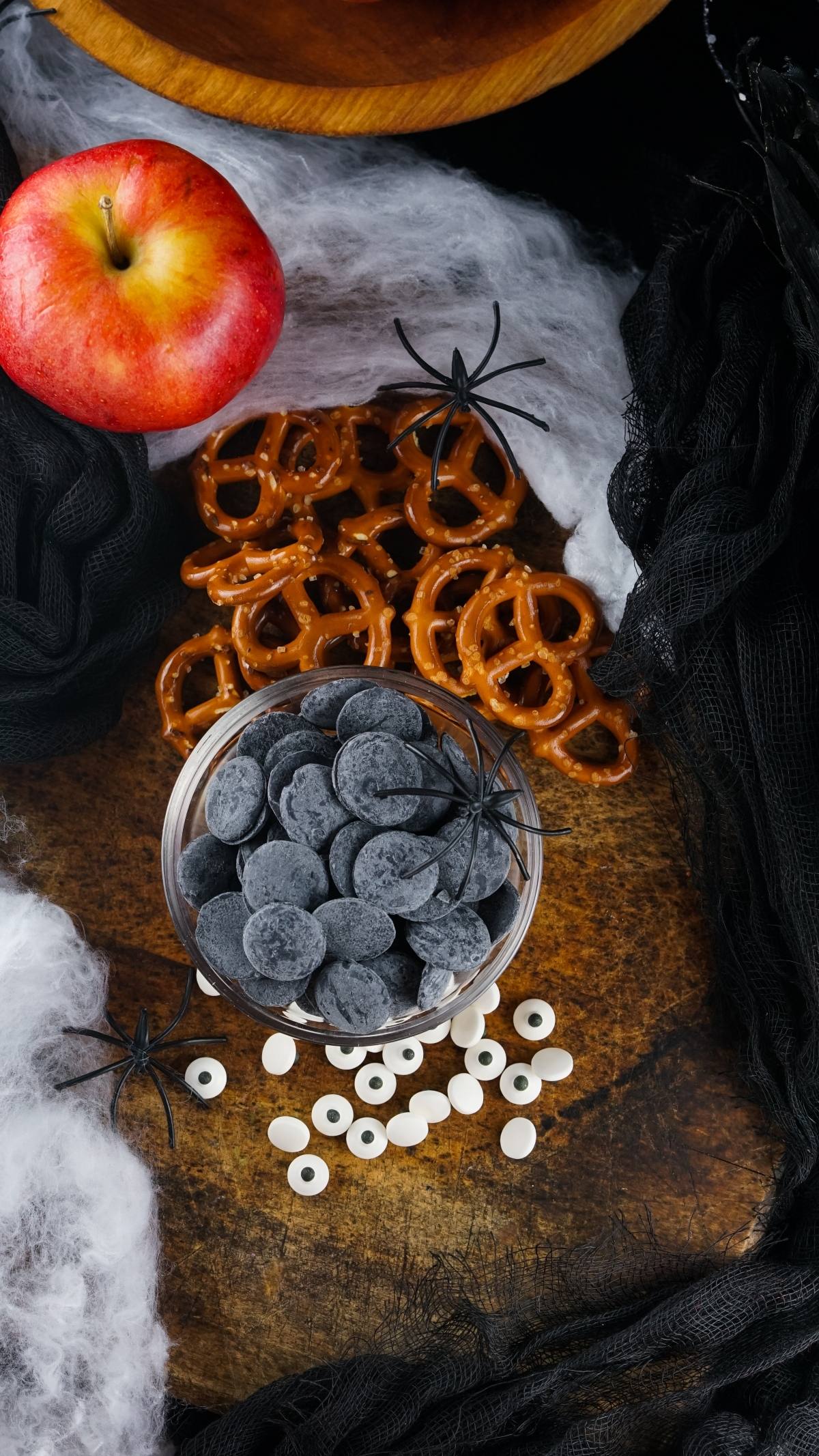 Spooky monster treats made with pretzels, black candies, candy eyes, apple slices, and fake spiders—an easy party treat recipe perfect as a Halloween snack for kids on a dark surface.