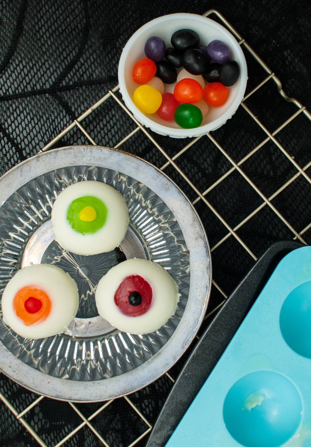 Three eyeball-shaped candies on a silver plate, with a bowl of colorful jellybeans nearby—perfect for gross looking Halloween food or creative Halloween party food ideas.
