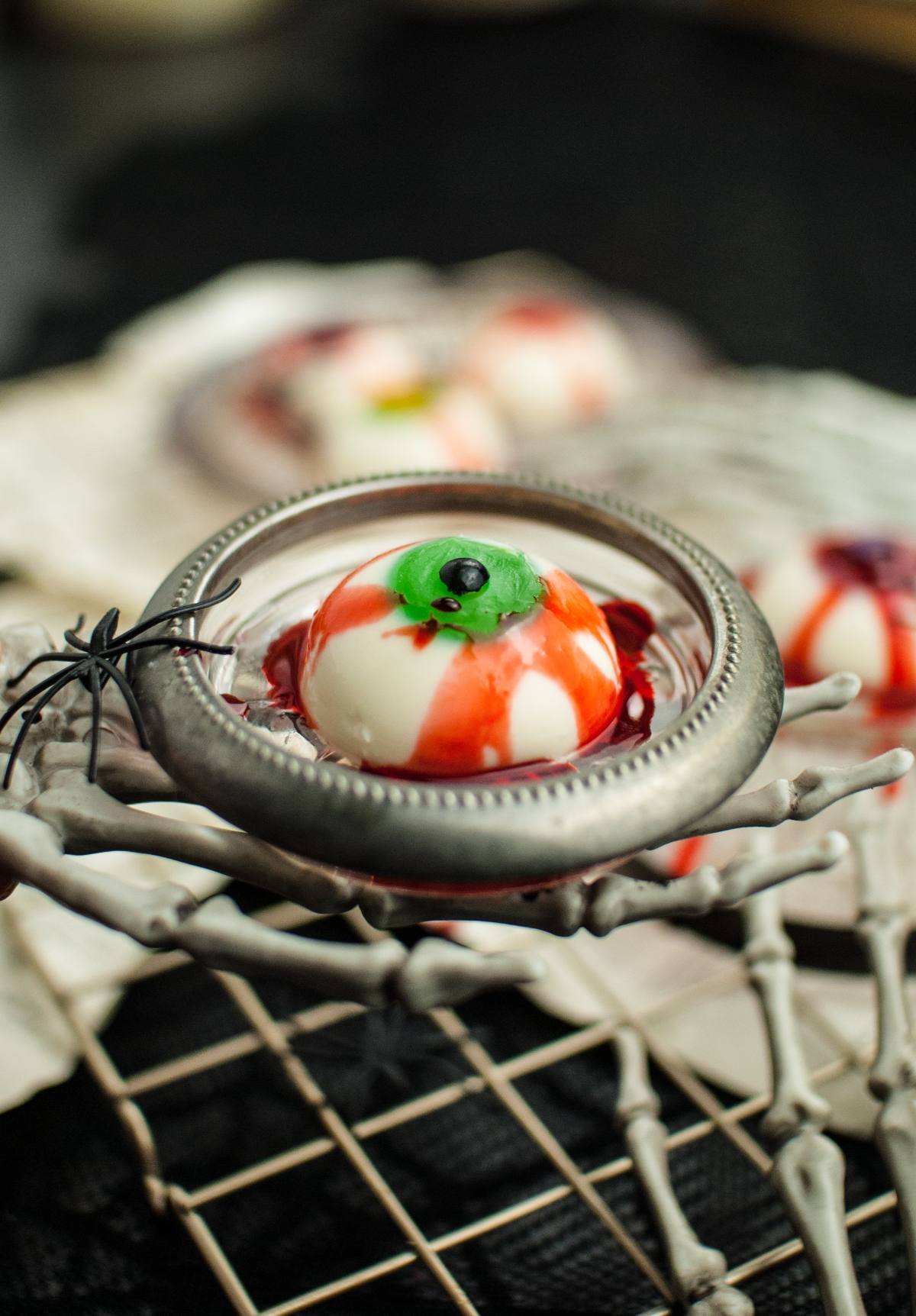 A spooky Gluten free Halloween dessert, this eerie Halloween Panna Cotta Eyeball sits on a metal dish held by a skeleton hand—perfect for your Halloween party food ideas.