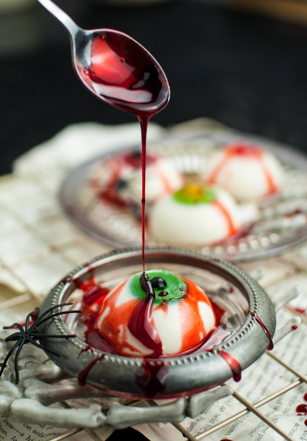 A spoon drizzles red syrup over a white dessert designed to look like a spooky eyeball—perfect for Halloween Panna Cotta Eyeballs or anyone craving gross looking halloween food.