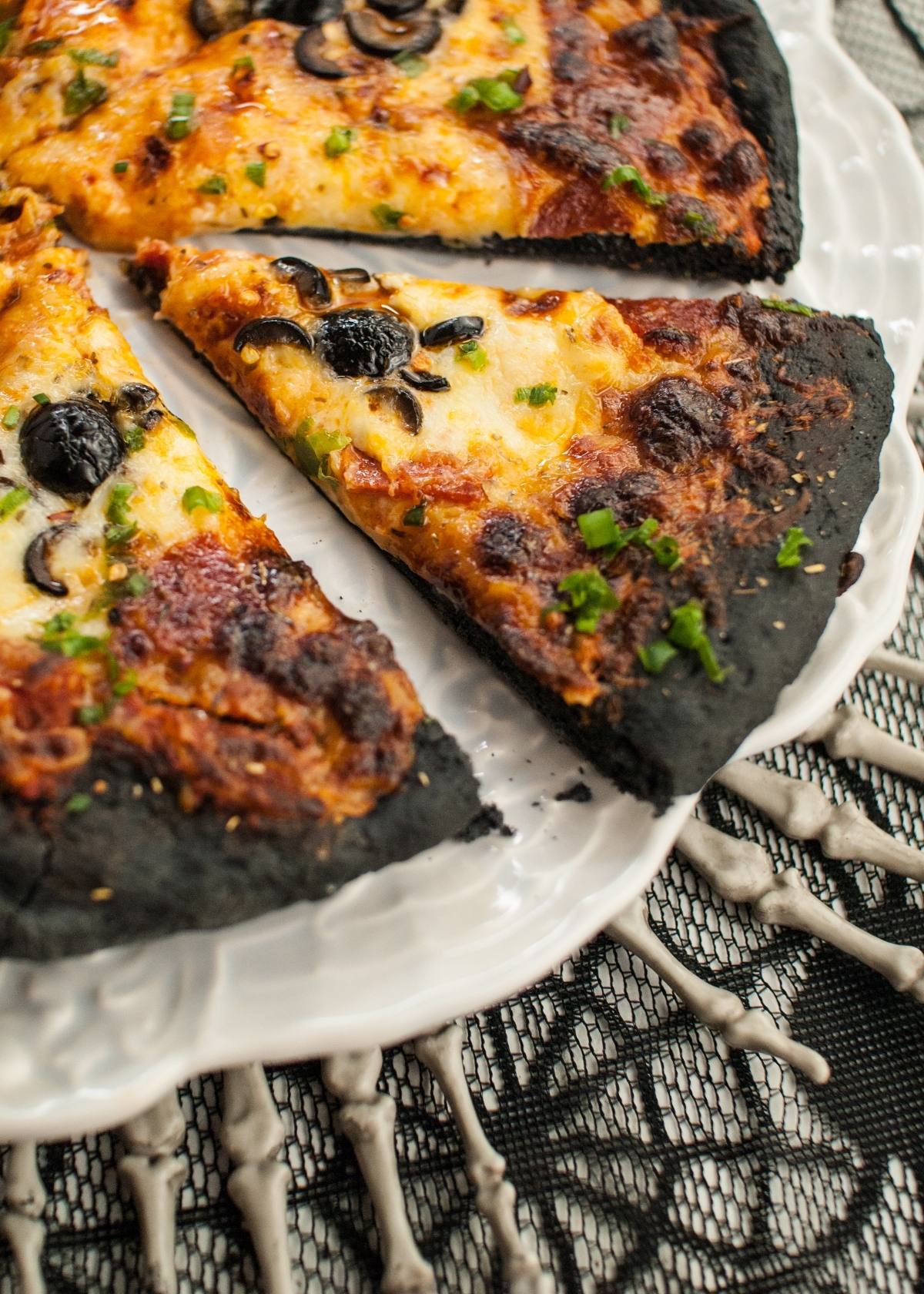 Slices of Halloween Pizza with Black Crust on a white plate, set atop a skeleton hand-patterned tablecloth—perfect for your Halloween dinner menu for adults and kids.