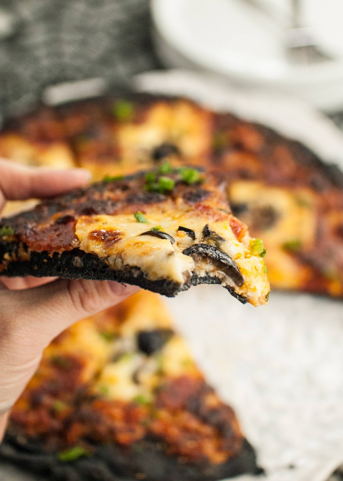 A hand holds a slice of pizza with melted cheese, mushrooms, and a blackened charcoal pizza dough—perfect for a Halloween Pizza with Black Crust.