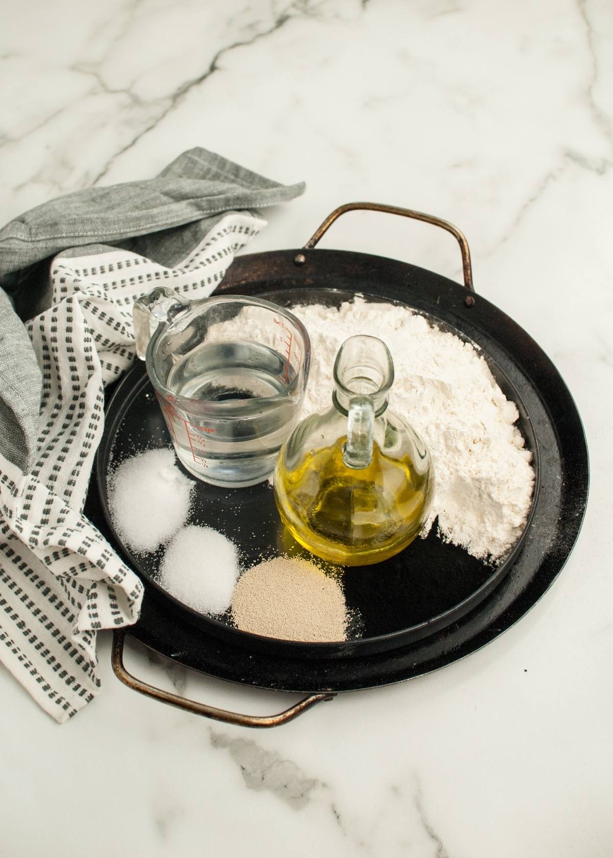 Baking tray with flour, olive oil, water, yeast, sugar, and salt on a marble surface—perfect for creating charcoal pizza dough for spooky Halloween pizza ideas that both adults and kids will love.