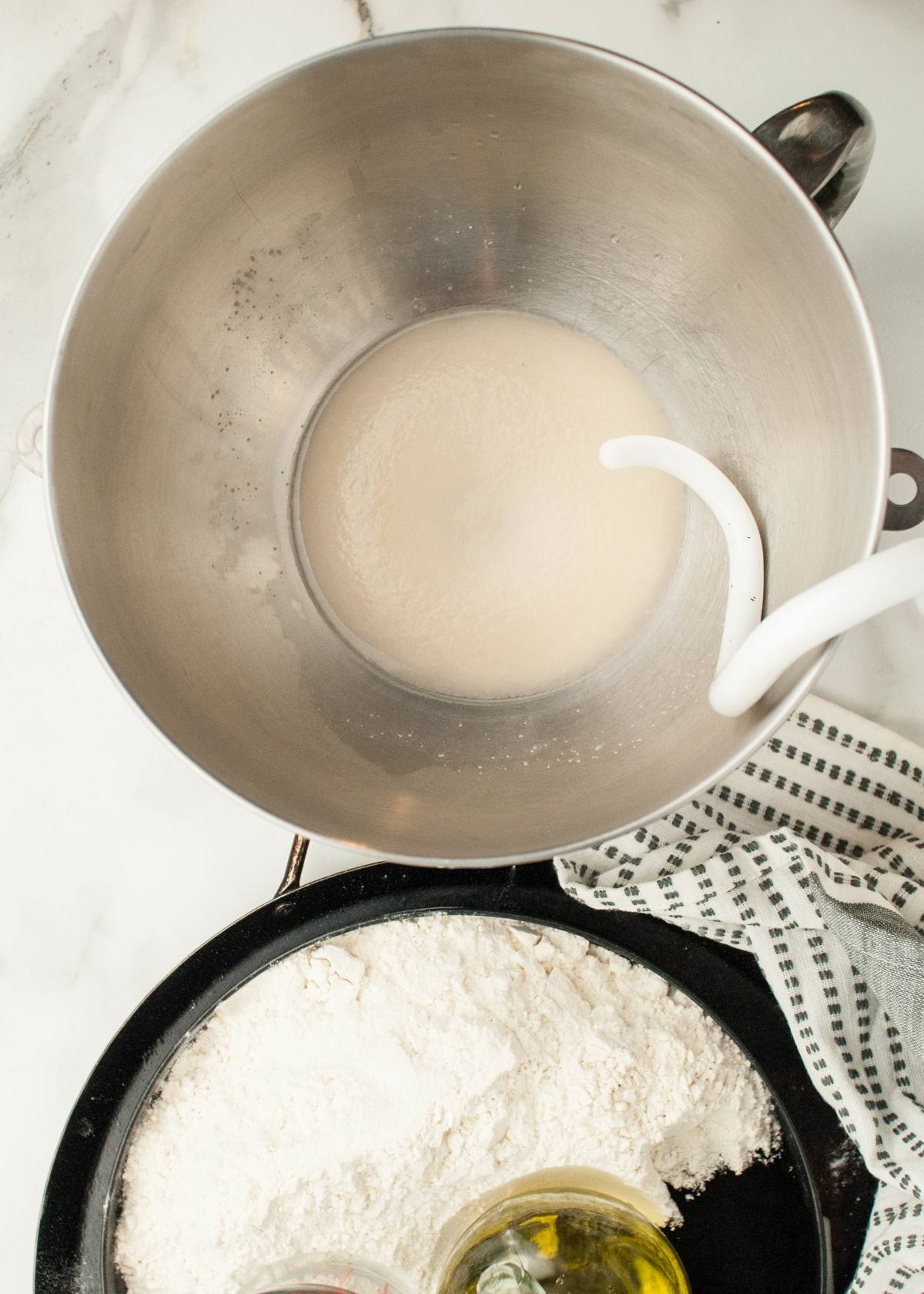 Metal mixing bowl with yeast mixture and dough hook beside a bowl of flour, oil, and a striped towel—perfect for prepping Halloween pizza ideas like pizza with black olive spiders.