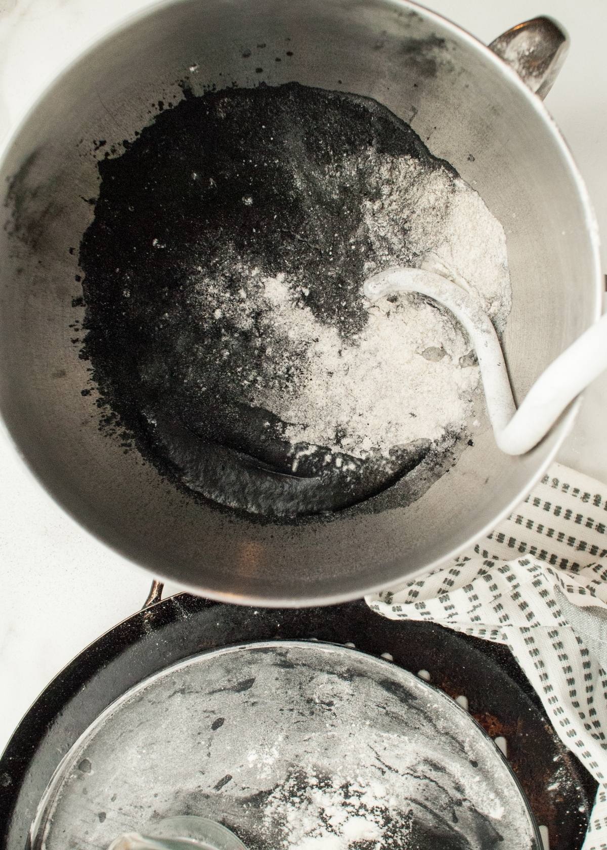 A mixing bowl with black and white flour mixture and a dough hook, next to a flour-dusted bowl and towel—perfect for creating Halloween pizza ideas like pizza with black olive spiders for your Halloween dinner menu for adults and kids.