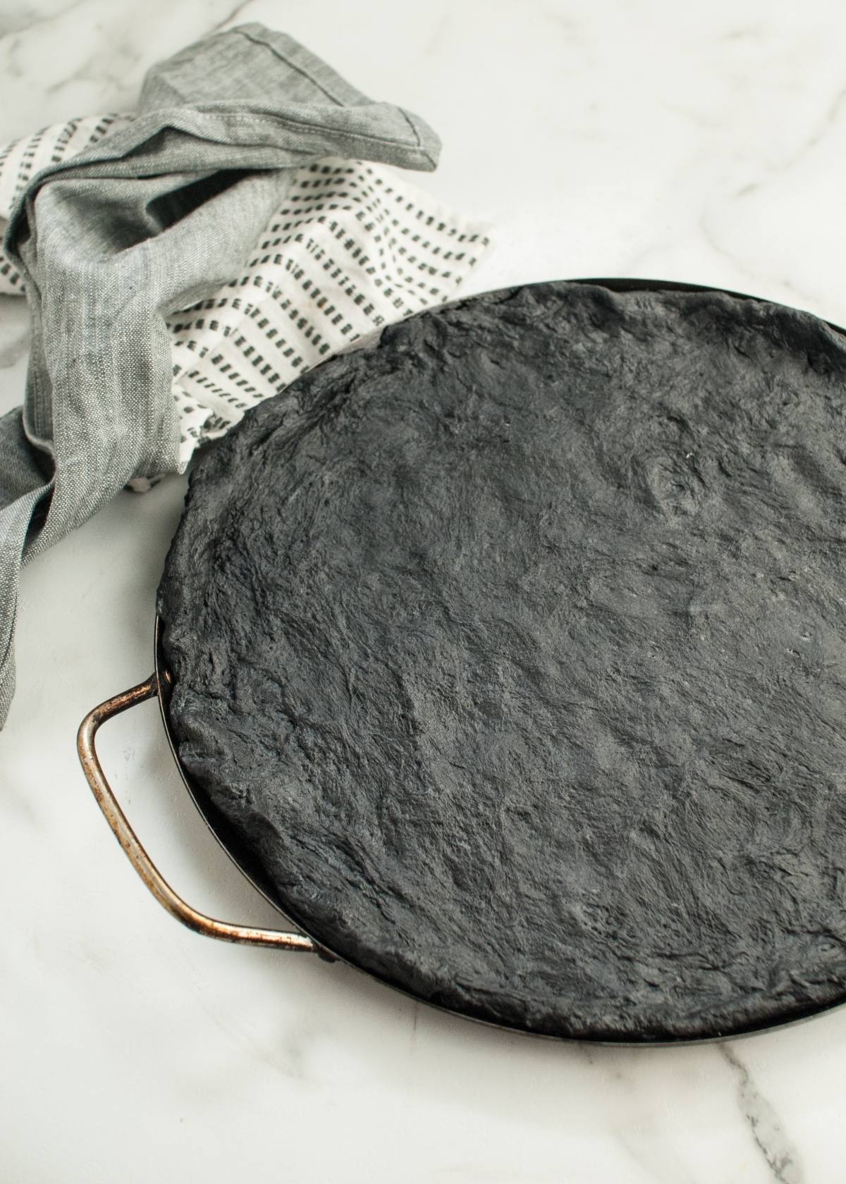 A Halloween Pizza with Black Crust sits on a round baking tray, its striking color perfect for spooky Halloween pizza ideas. A kitchen towel rests nearby on the white marble surface.