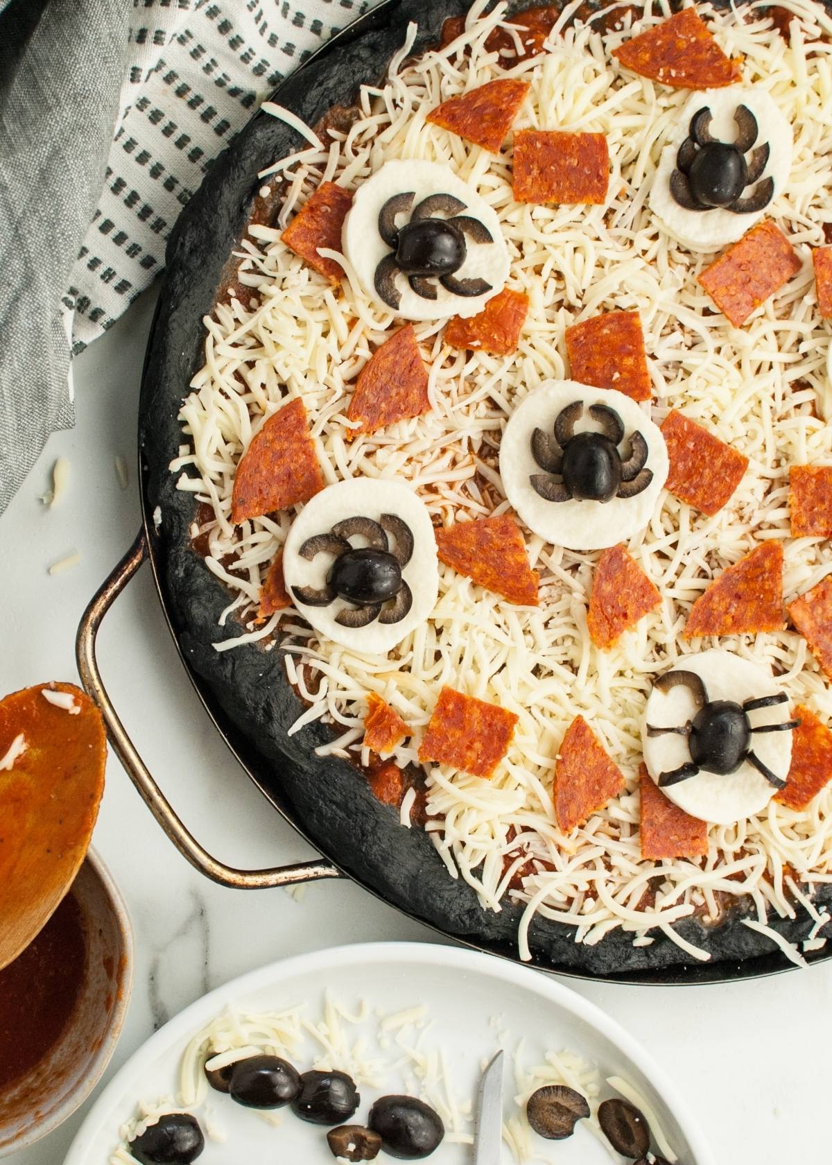 Unbaked Halloween Pizza with Black Crust, topped with mozzarella, pepperoni, and cute black olive spiders on onion slices—perfect for a spooky Halloween dinner menu for adults and kids.