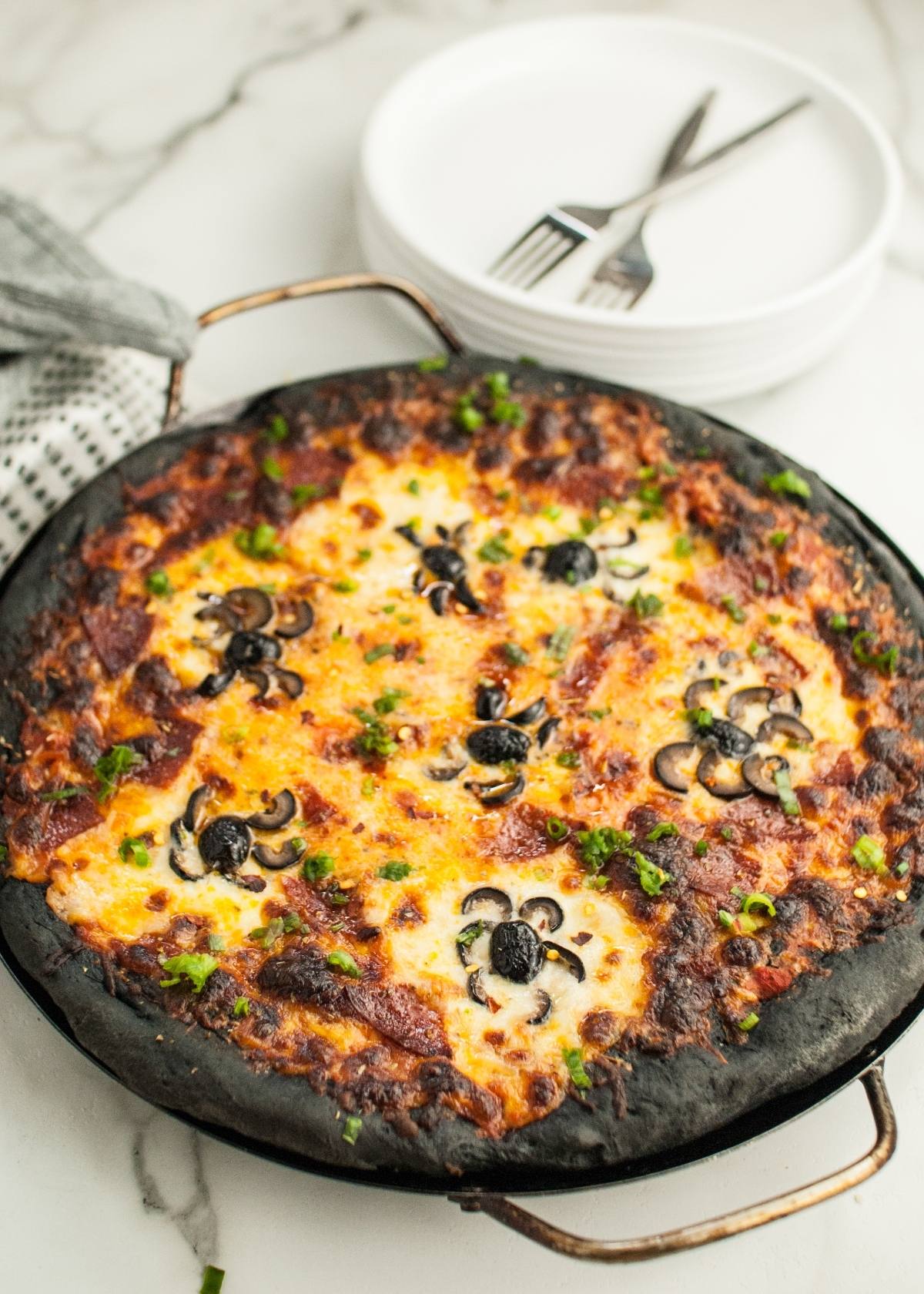 A Halloween Pizza with Black Crust, topped with cheese, green herbs, and pizza with black olive spiders, sits on a pan beside stacked plates and forks.