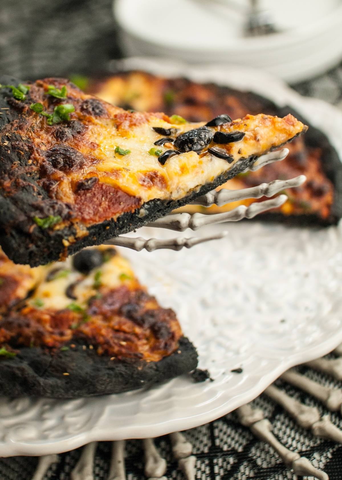 A slice of pizza with charcoal pizza dough and black olive spiders, served on a skeleton hand plate—perfect for spooky Halloween pizza ideas.