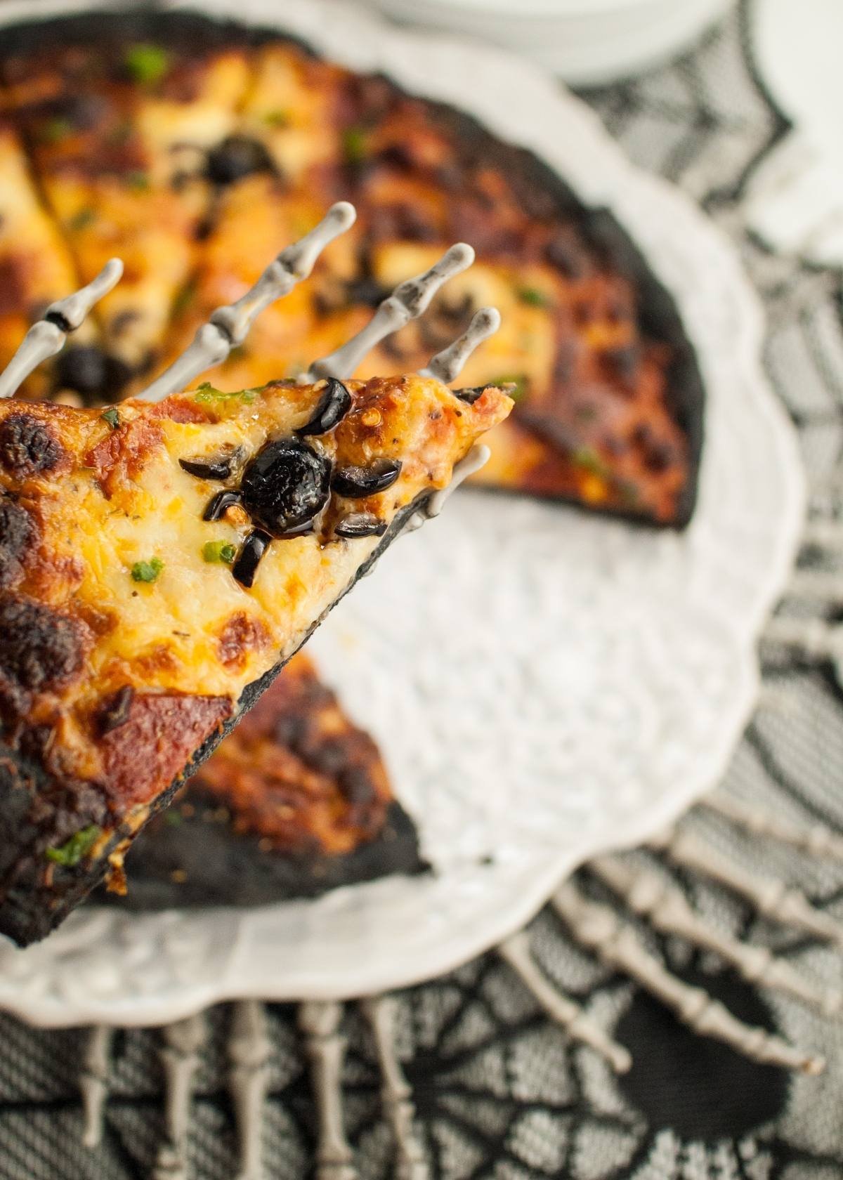 A skeleton hand holds a slice of pizza with black olive spiders on a spiderweb-patterned tablecloth—perfect for spooky Halloween pizza ideas.