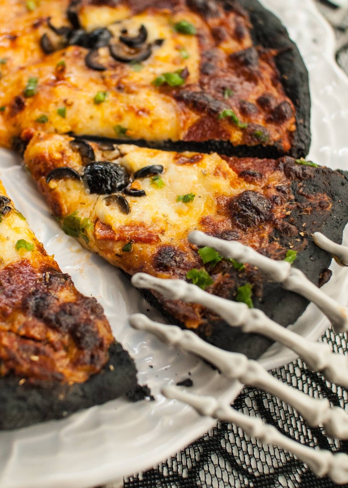 A skeleton hand serving a slice of charcoal pizza dough topped with cheese, olives, and herbs—a spooky Halloween dinner menu for adults and kids.
