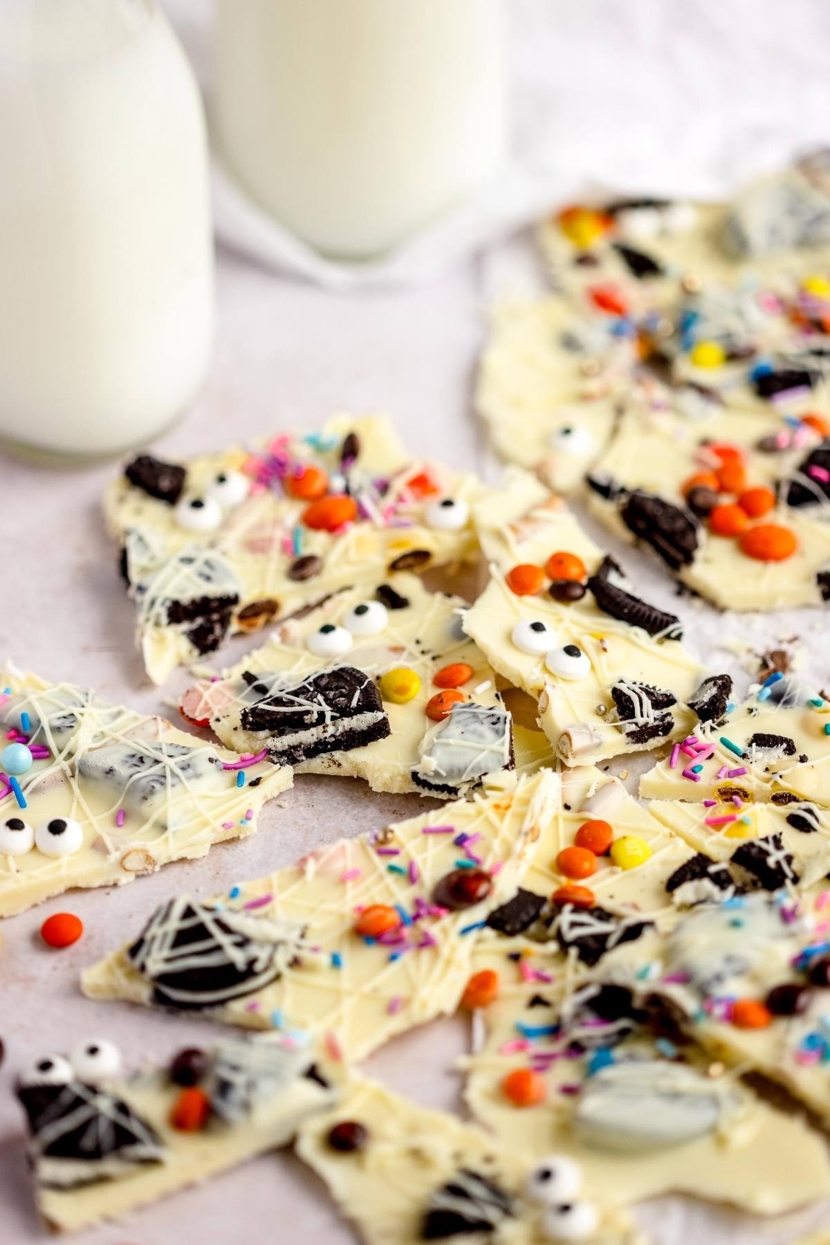Colorful Halloween-themed white chocolate bark with candy eyes, sprinkles, and Oreo pieces—an easy candy bark recipe for a fun Halloween treat with googly monster eyes, served next to bottles of milk.